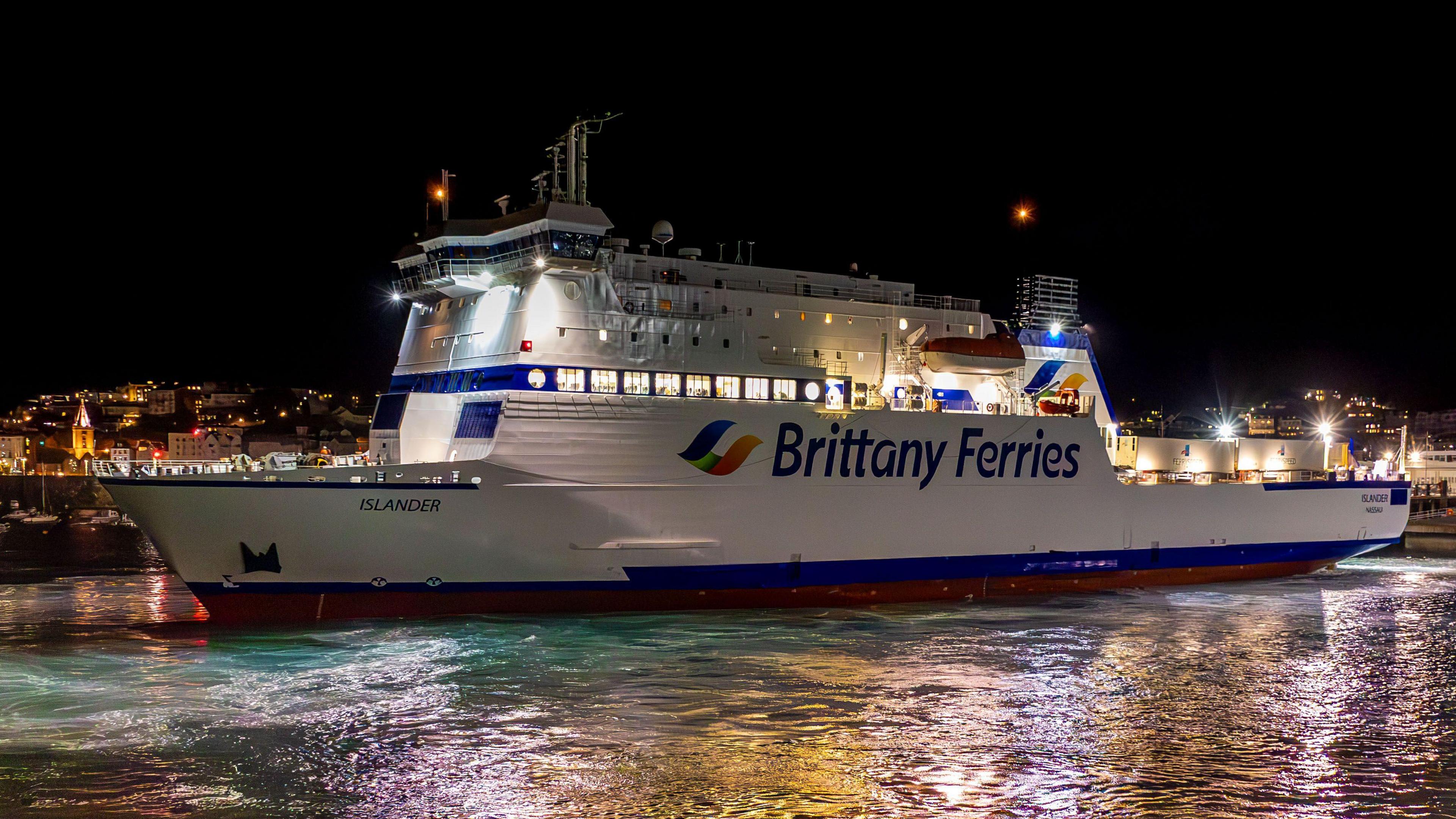 A Brittany Ferries boat called Islander docked in St Peter Port at night. Lights from the town can be seen behind the ferry. A number of lights are on the boat.