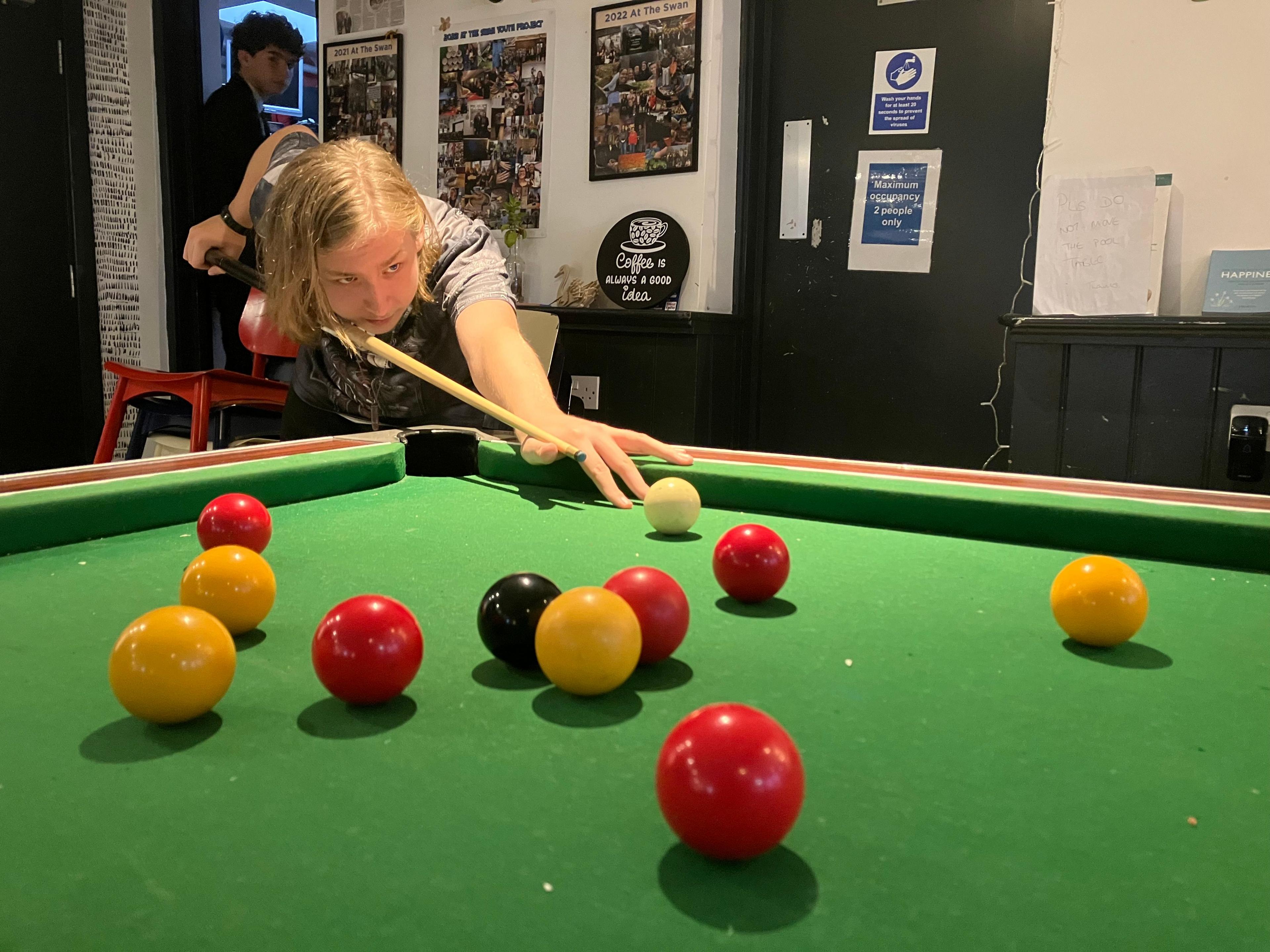 A picture of a teenage boy with long blonde hair who is playing pool.