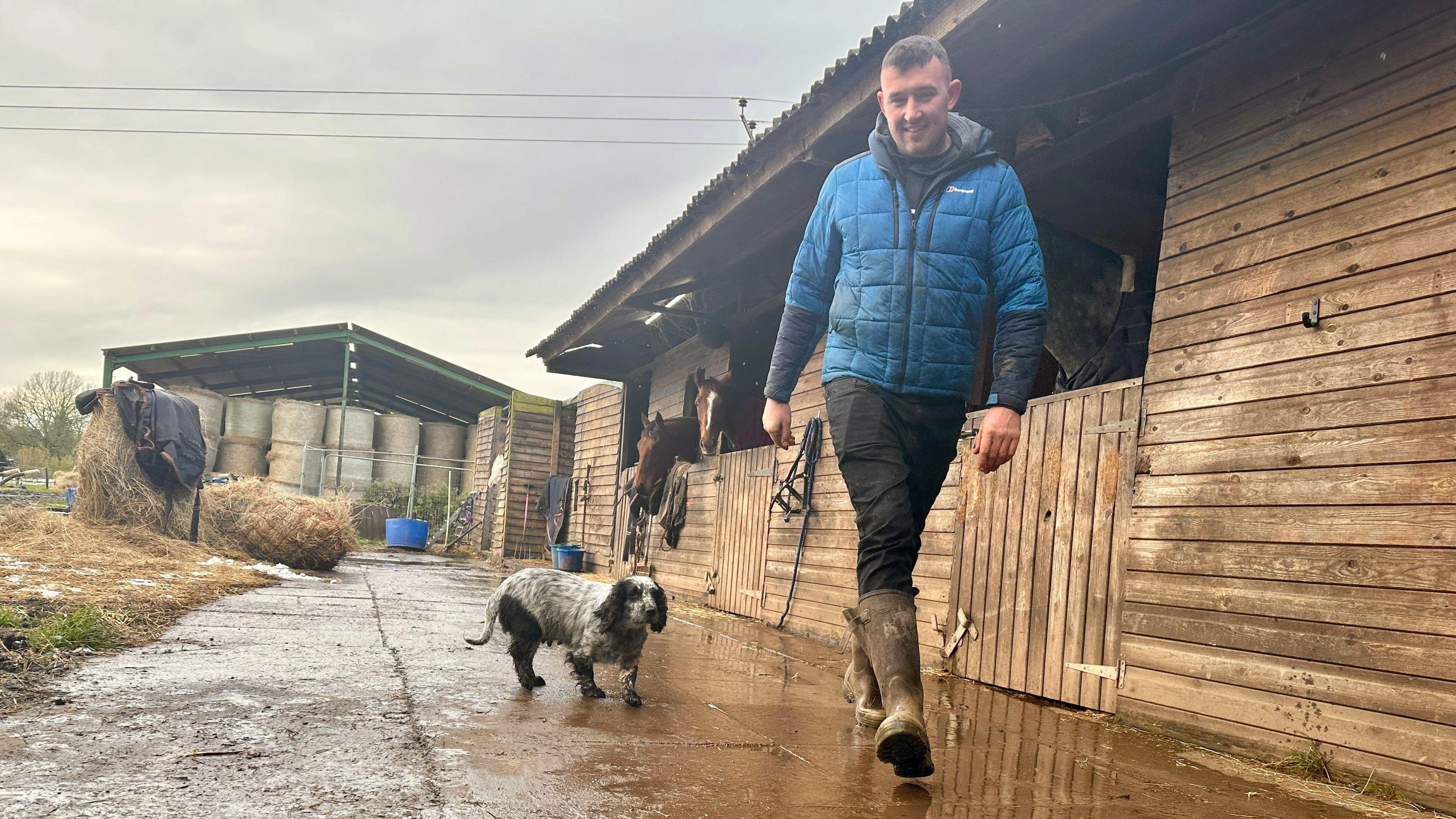 Farmer Andrew Jennings wears a blue jacket, black trousers and wellington boots as he walks through a farmyard next to stables, accompanied by a small black and white dog.