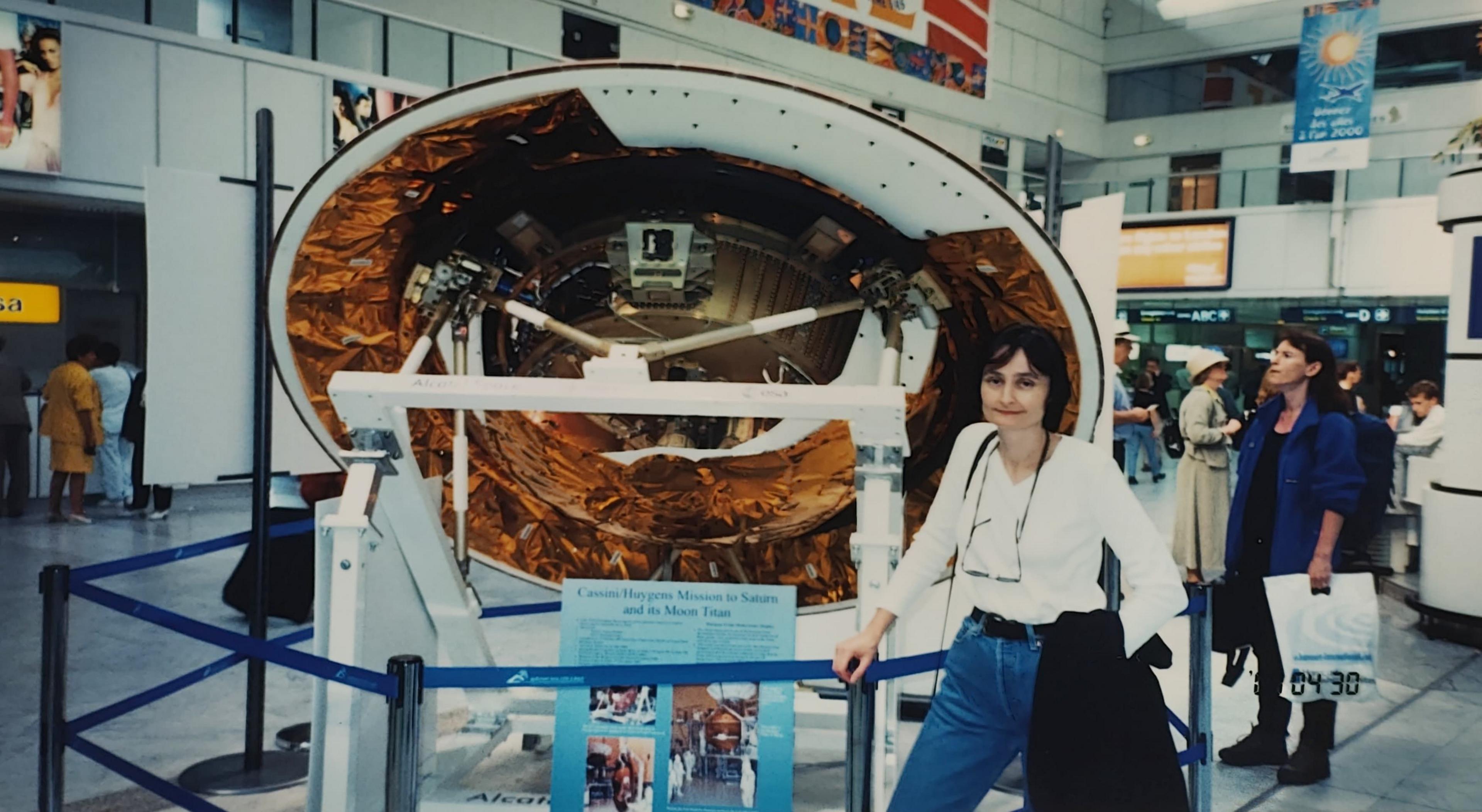 Michele Dougherty age 30 in a white top and blue jeans with glasses hanging around her neck and a black jacket slung over her arm. She stands in front of a round part of a spacecraft which is partly white but with copper coloured foil in parts. It is cordoned off by a blue ribbon rail. The scene appears to be in a museum, or perhaps and airport and other people are milling around.