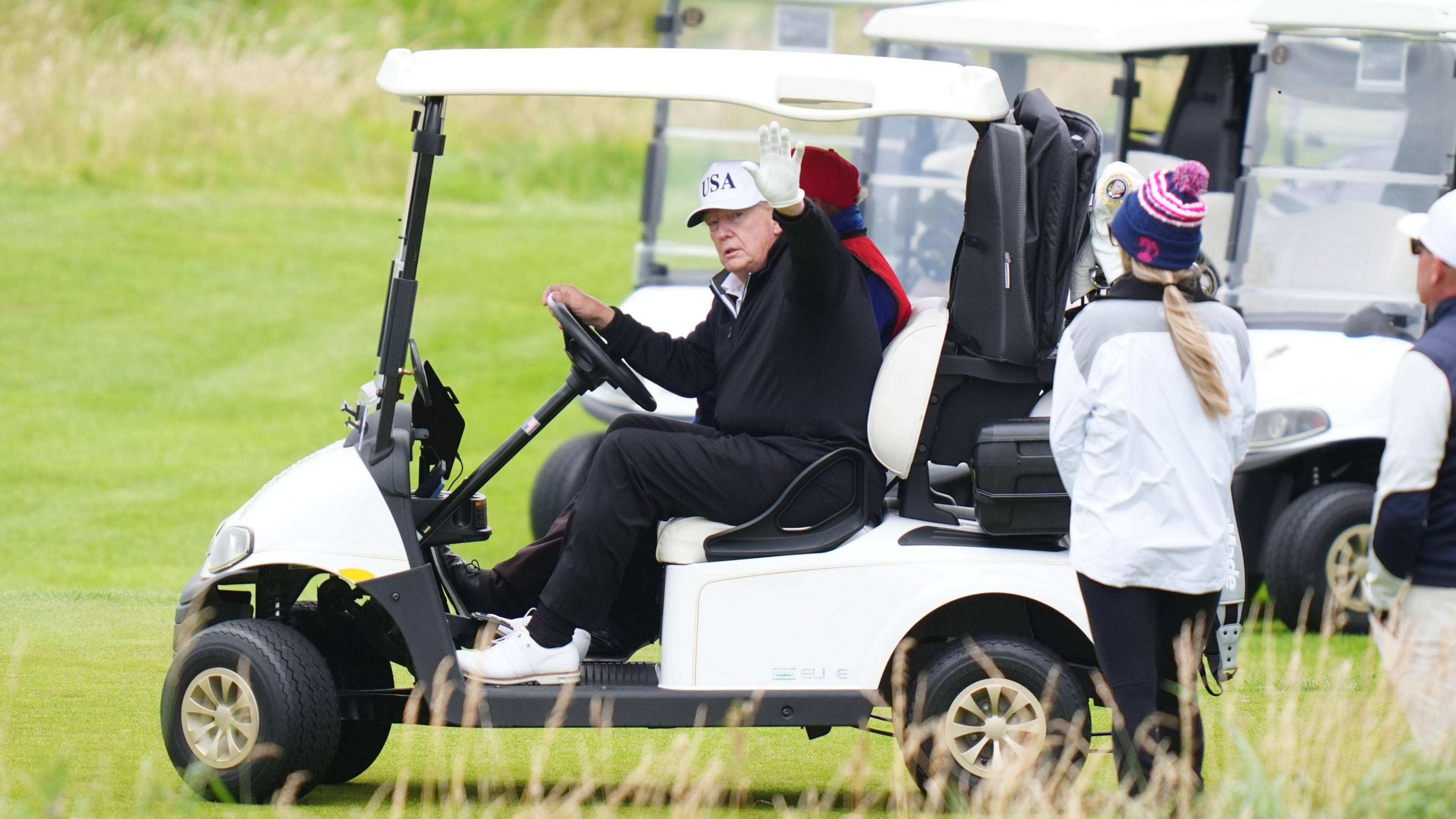 Donald Trump waves from a golf buggy while wearing dark-coloured waterproof golf clothes and a white cap. A woman wearing white waterproof jacket and blue wooly hat stands next to him