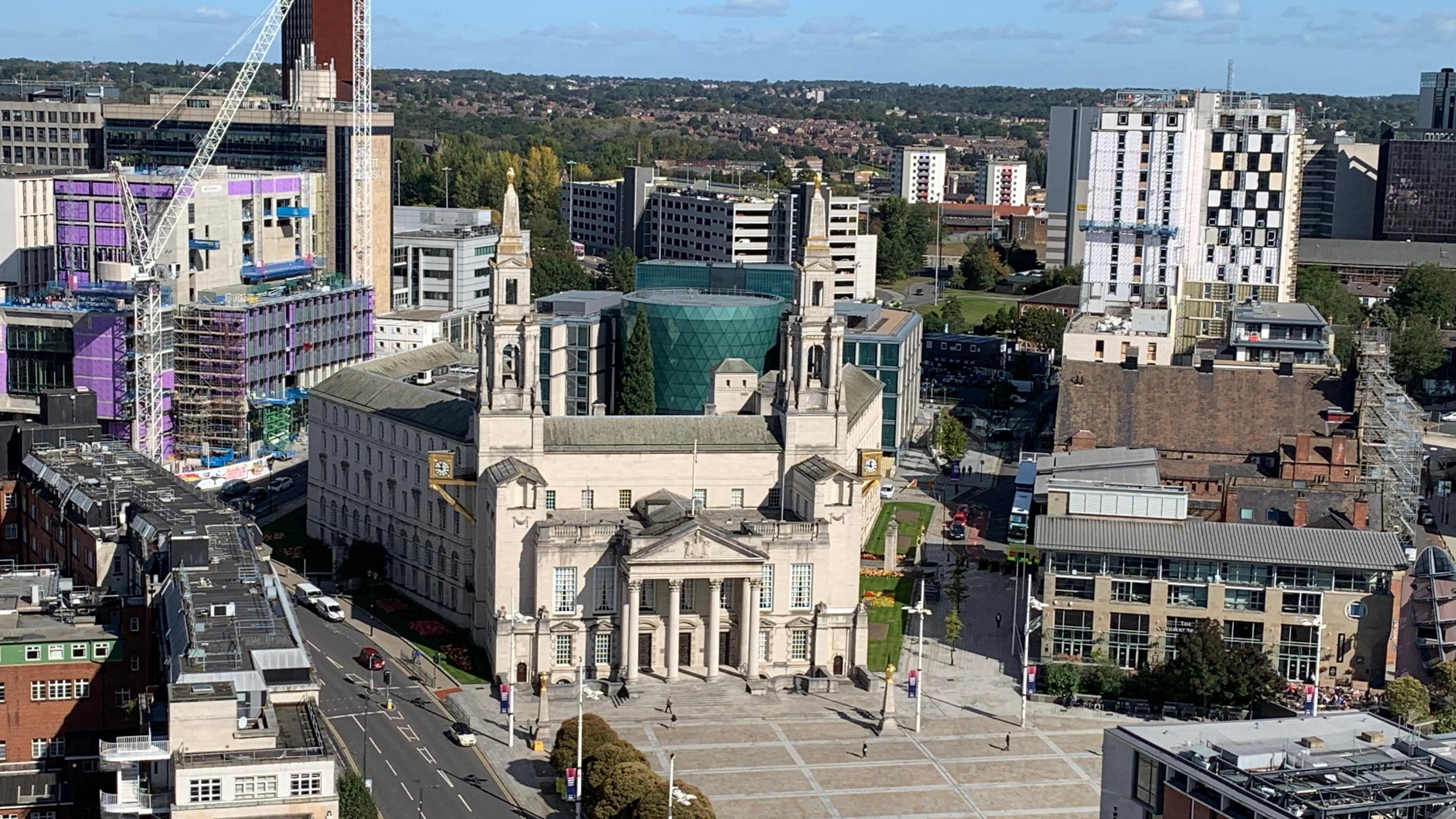 Image shows an ariel shot of Leeds Civic Hall with Millenium Square.
Behind is a Leeds University glass building with a sky line scene.
To the left of the shot is the Leeds General Infirmary.