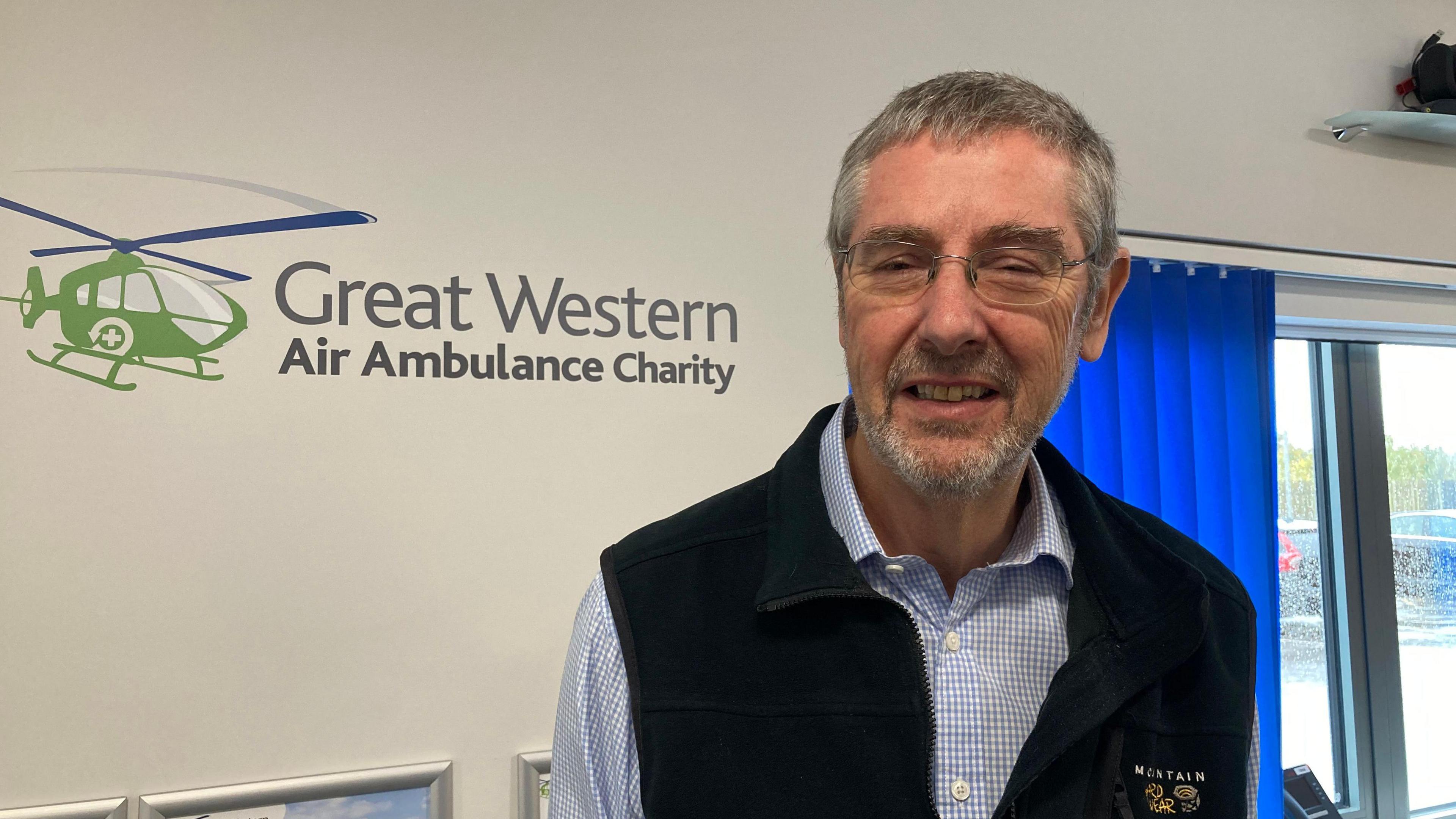 Forrest Wheeler smiles at the camera at the Great Western Ambulance Charity, he is wearing a white and blue checked shirt with a navy gilet.