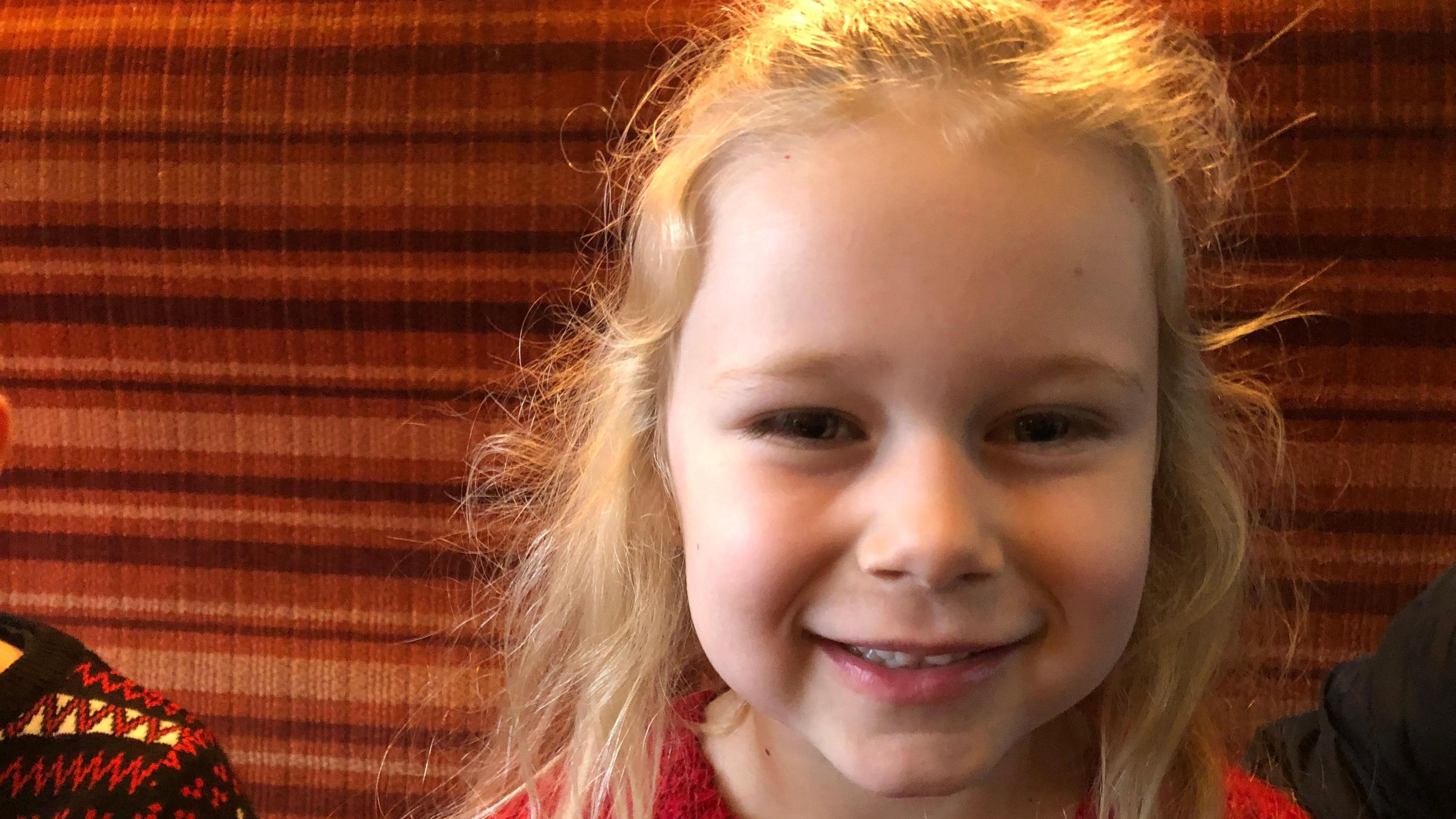 Six-year-old Violet has blonde hair over her shoulders and is wearing a red woollen jumper. She is smiling as she sits in a railway carriage