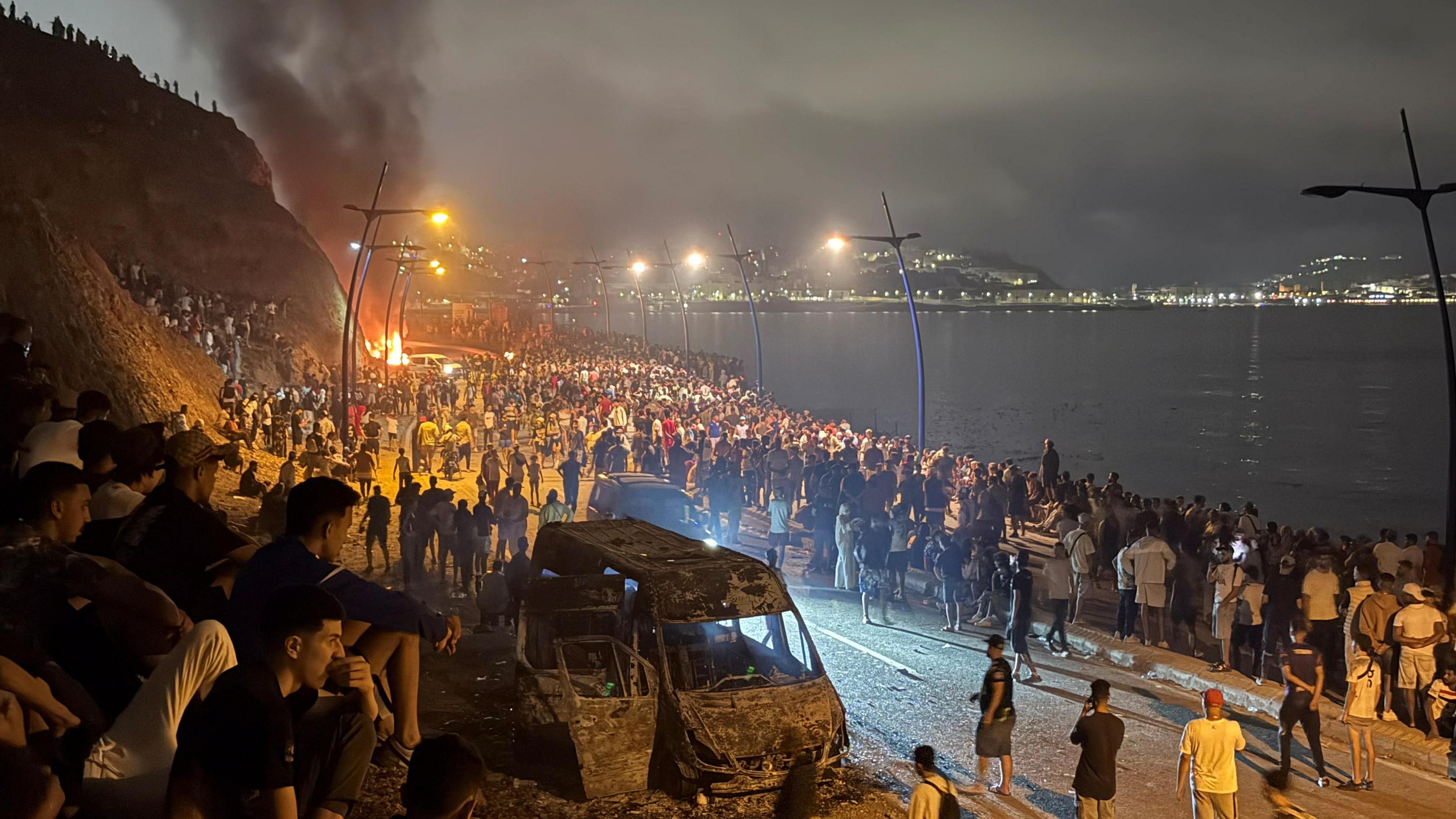 Crowds gather around a torched vehicle while waiting for a chance to cross to the Spanish enclave of Ceuta, on the Moroccan side of the border in Fnideq, Morocco