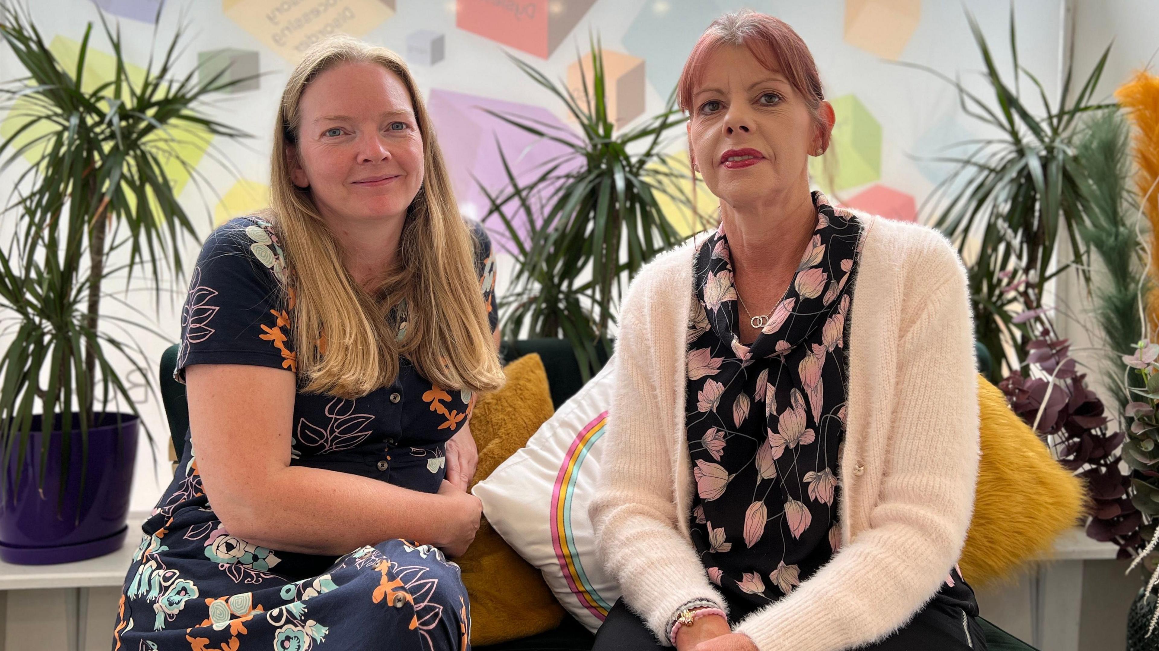 Niamh and Penny are sitting on a sofa surrounded by plants. Niamh is wearing a print dress and Penny has a print shirt with a pink fleece over the top.