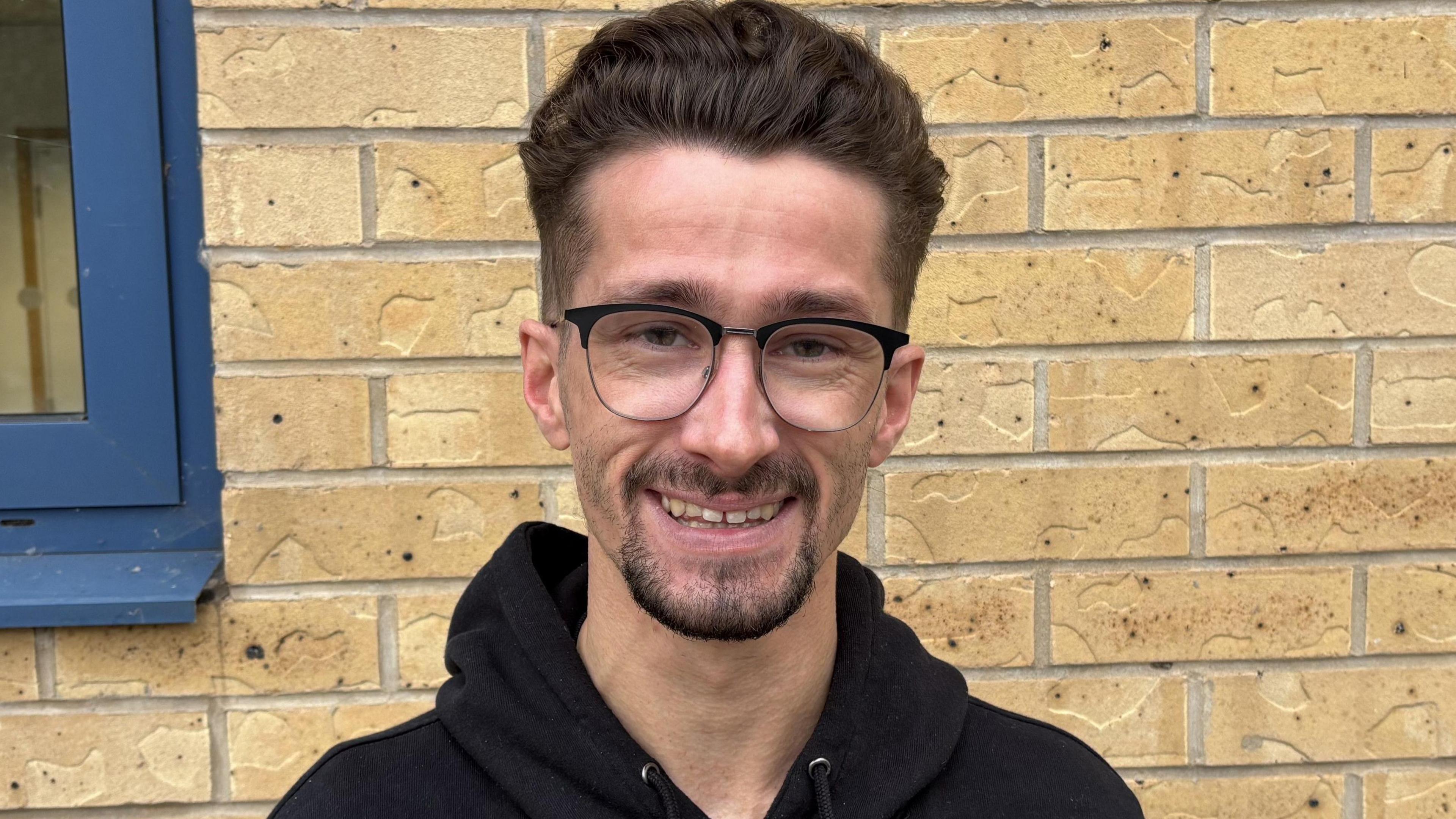 A headshot of a man standing in front of a bright wall. He has thick brown hair which is styled upwards and short on the sites. He has a goatee and wears thin-rimmed glasses. He is wearing a black hoodie.