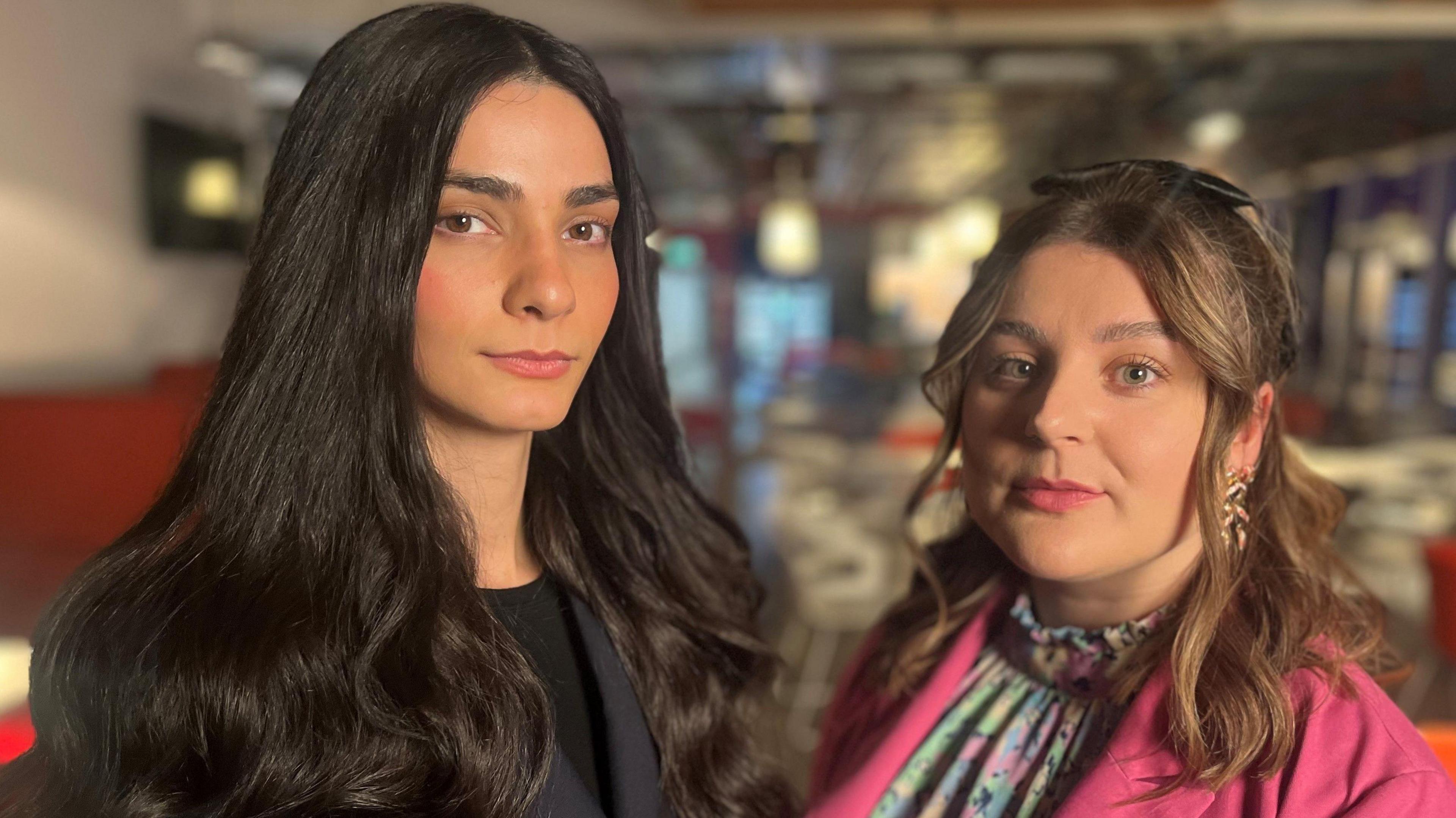 Ellie Wilson & Hannah McLaughlan are photographed looking directly at the camera. The background is blurred out but it is the BBC Scotland newsroom.    Ellie has long dark hair and is wearing a dark suit. Ellie is smaller, with shoulder-length hair and a side-parting. She is wearing a pink jacket and has a colourful shirt. 