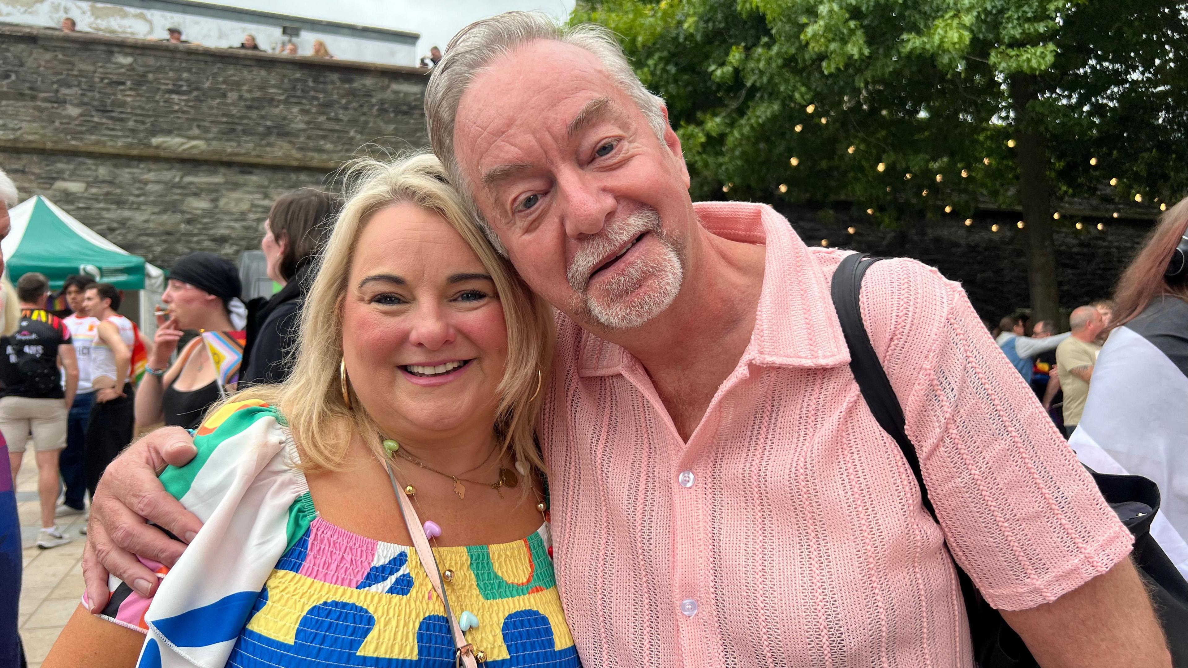 A woman wearing a colourful top smiles at the camera. Next to her is a man in a short-sleeved pink shirt. He has his right arm around the woman's shoulder.