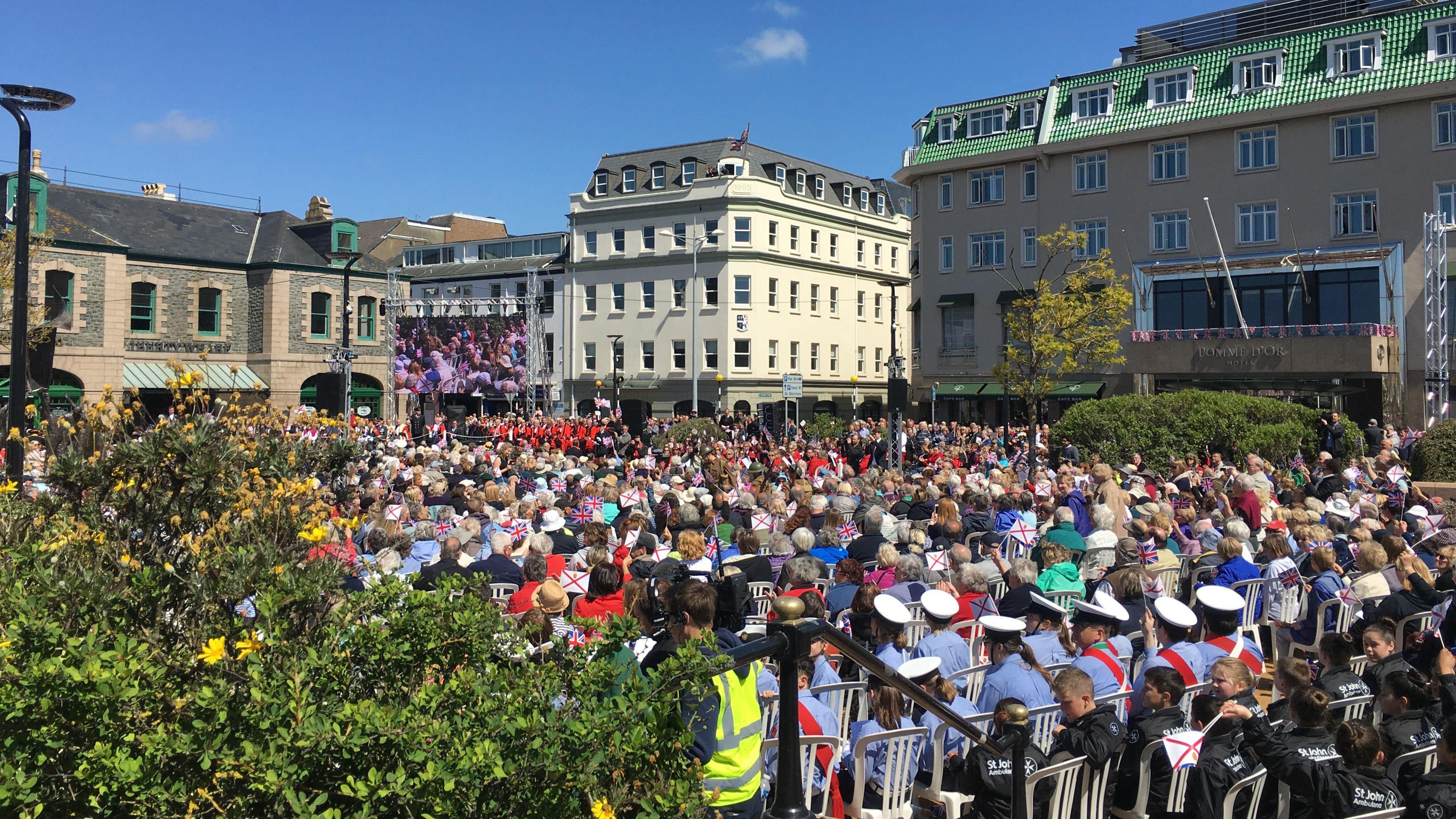 A crowd of people sitting in Liberation Square in St Helier on a sunny day to celebration Liberation Day. There are about 100 people in the picture.