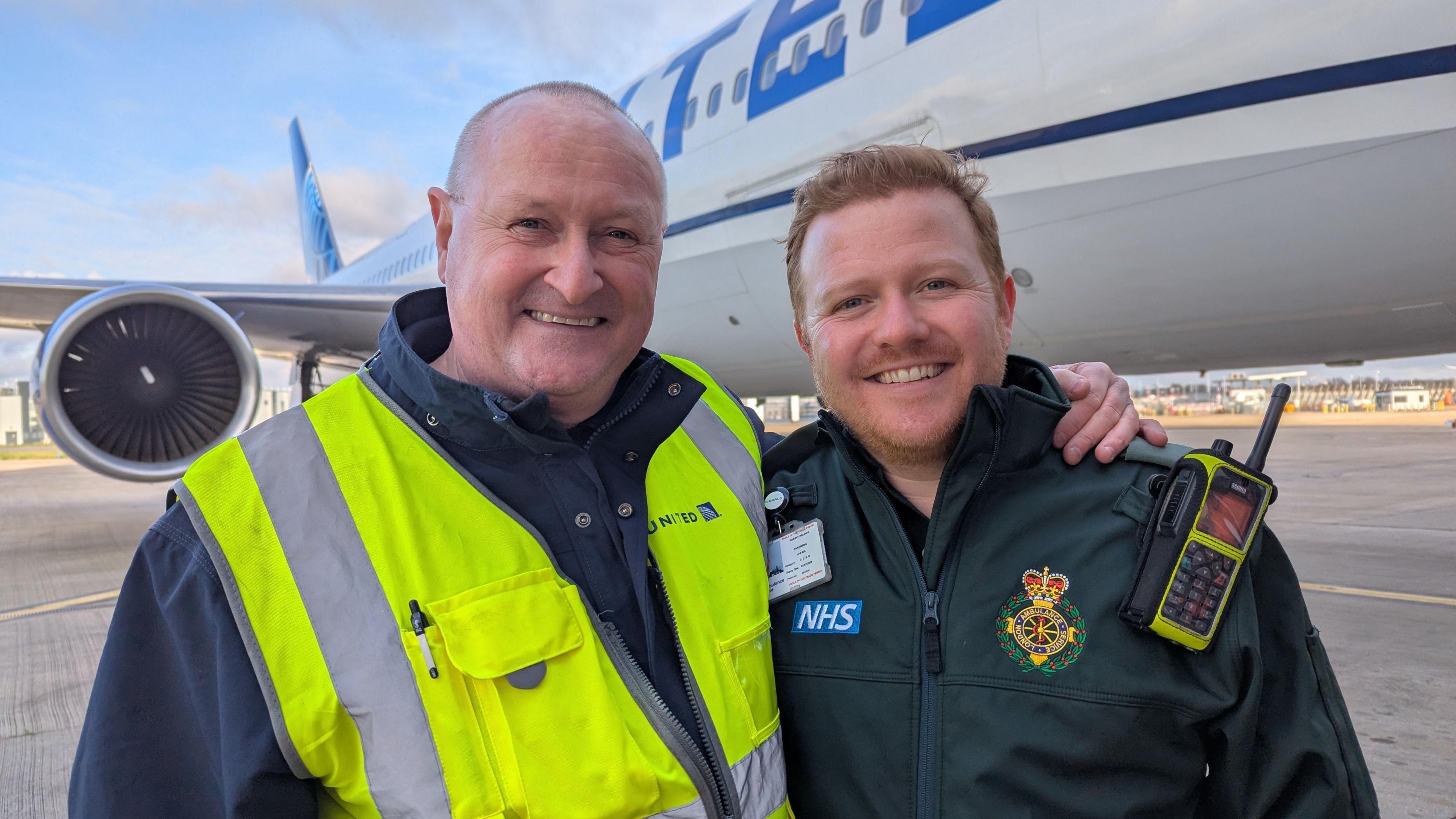 John Walton (left) stands with his arms around paramedic Rob Weldon. Mr Walton is wearing a navy blue jacket with an illuminous vest on top while Mr Weldon is wearing a paramedic uniform, complete with a walkie-talkie on his left shoulder. They are standing in front of a plane at an airport.