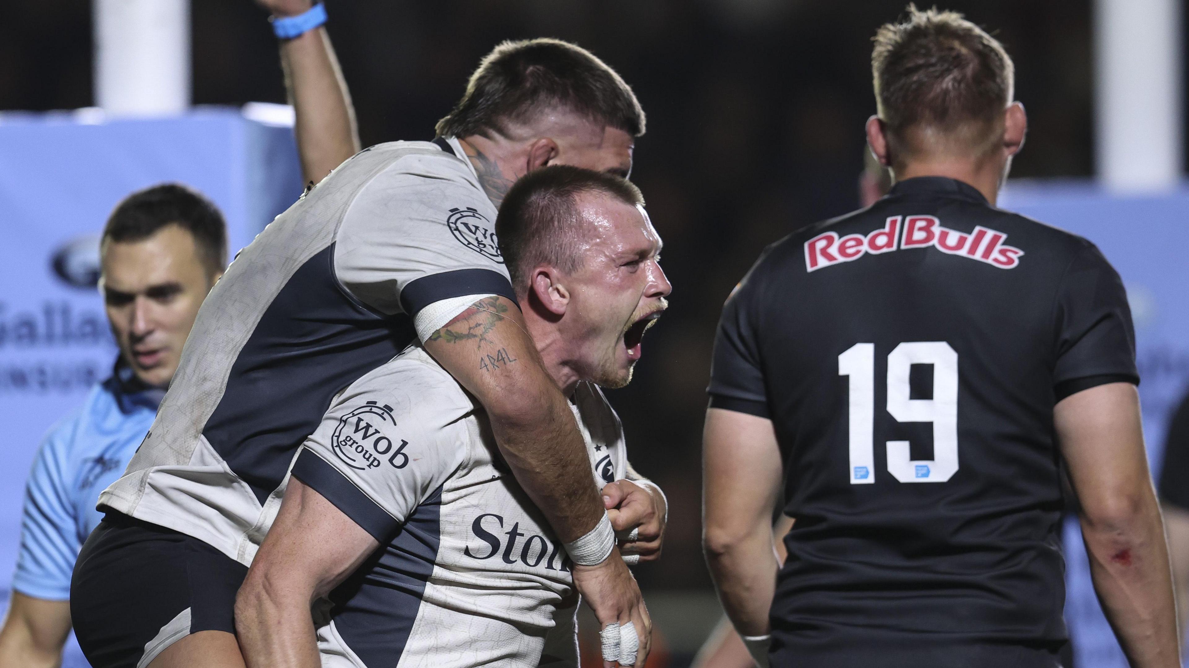 Nick Tompkins of Saracens roars with delight after scoring a try as Theo Dan jumps on his back with Newcastle no.19 Freddie Clarke having his back turned
