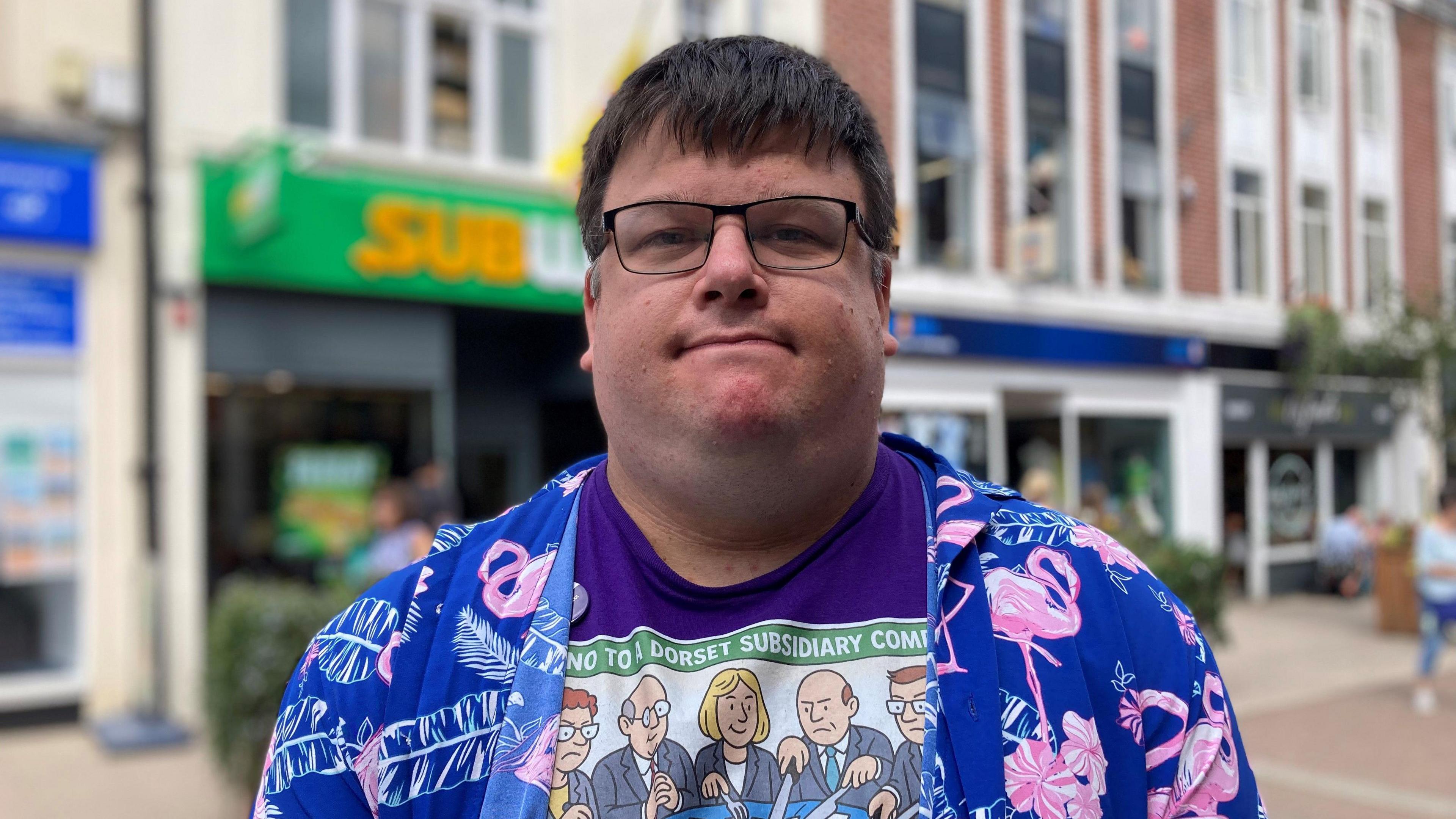 A head-and-shoulders photo of Frank Goodwin, who has short brown hair, rectangular glasses, and is wearing a purple Unison T-shirt with a blue and pink patterned shirt over the top. A row of shops can be seen behind him.