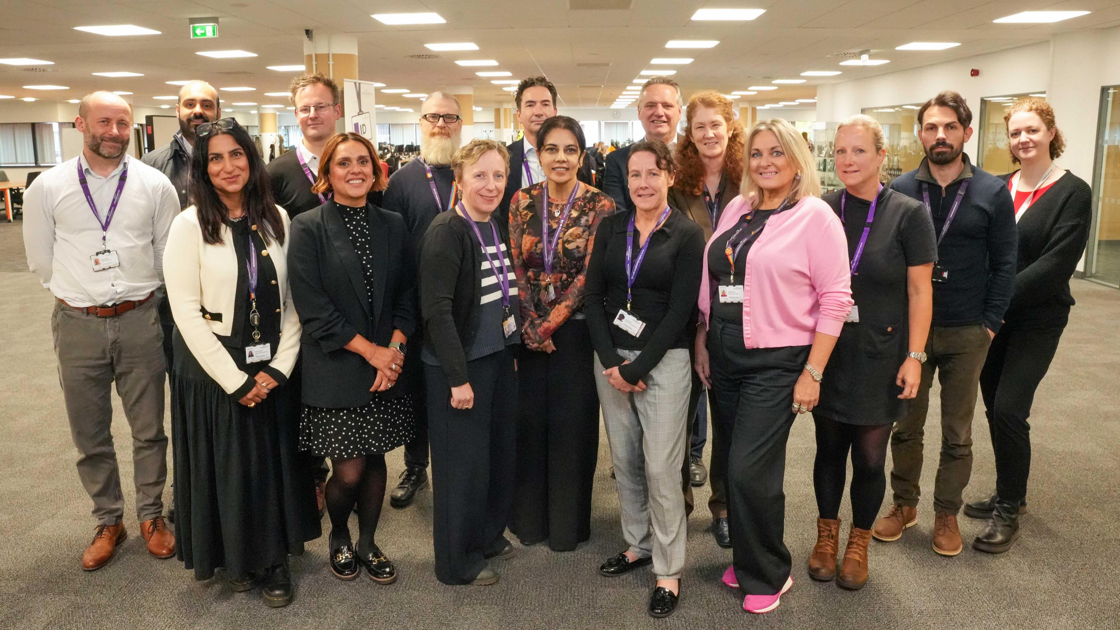 A group of adults gathered in an open conference room. They are smiling and wearing purple lanyards. They are all dressed in office wear.
