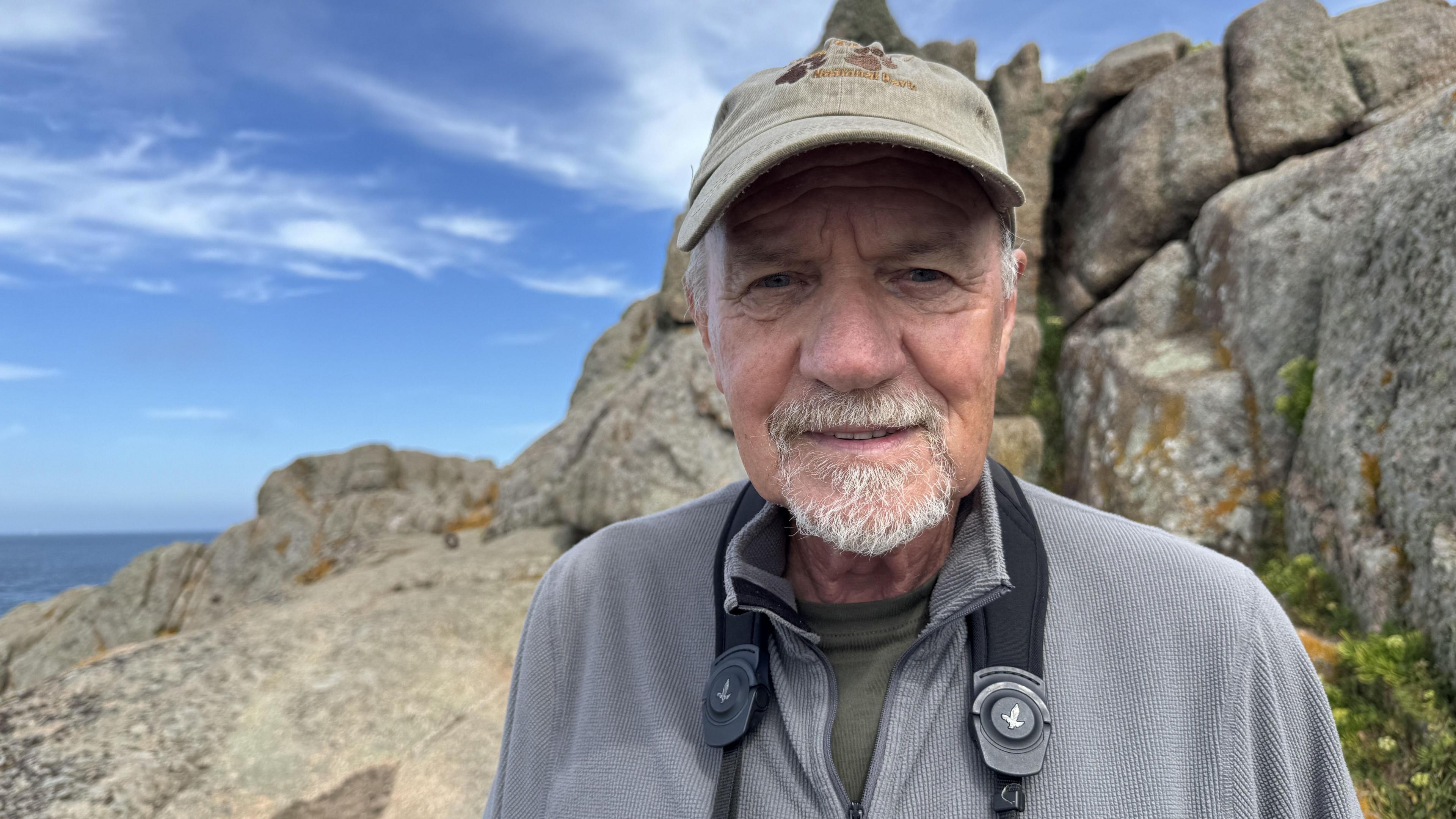A man stands in a rugged outdoor setting beneath a bright blue sky dotted with light clouds. He's wearing a grey jacket and cap and has binoculars hanging from his neck. 
