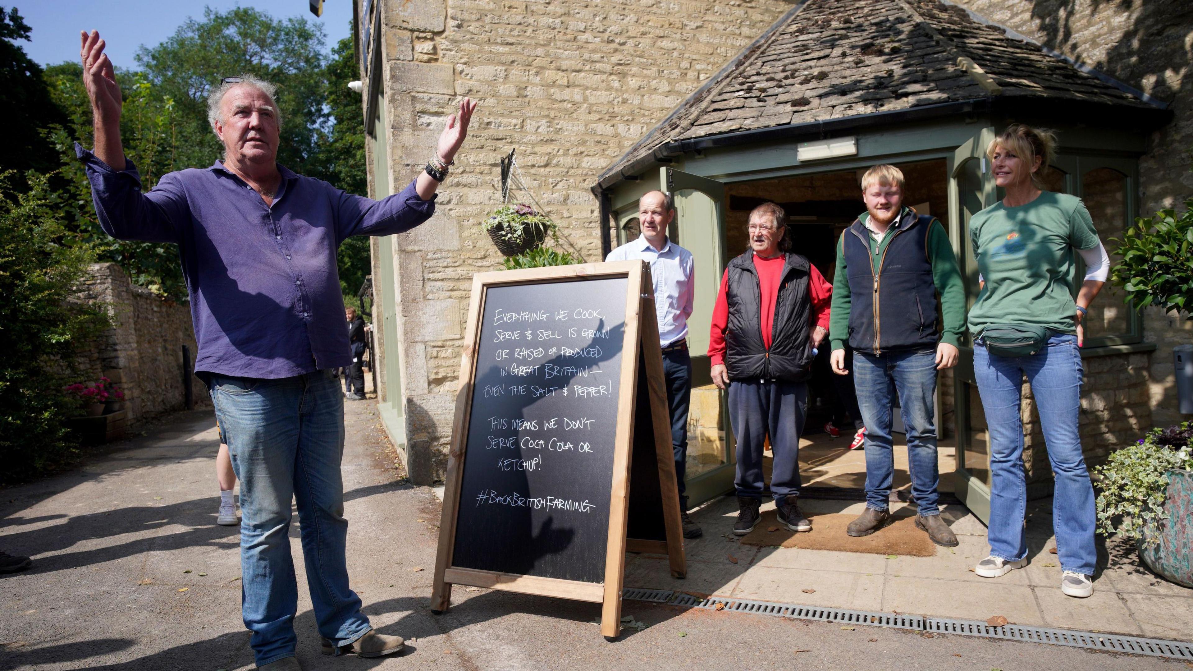 Jeremy Clarkson on the left outside the pub next to a blackboard sign. To the right there are four people stood outside the front door of The Farmer's Dog.