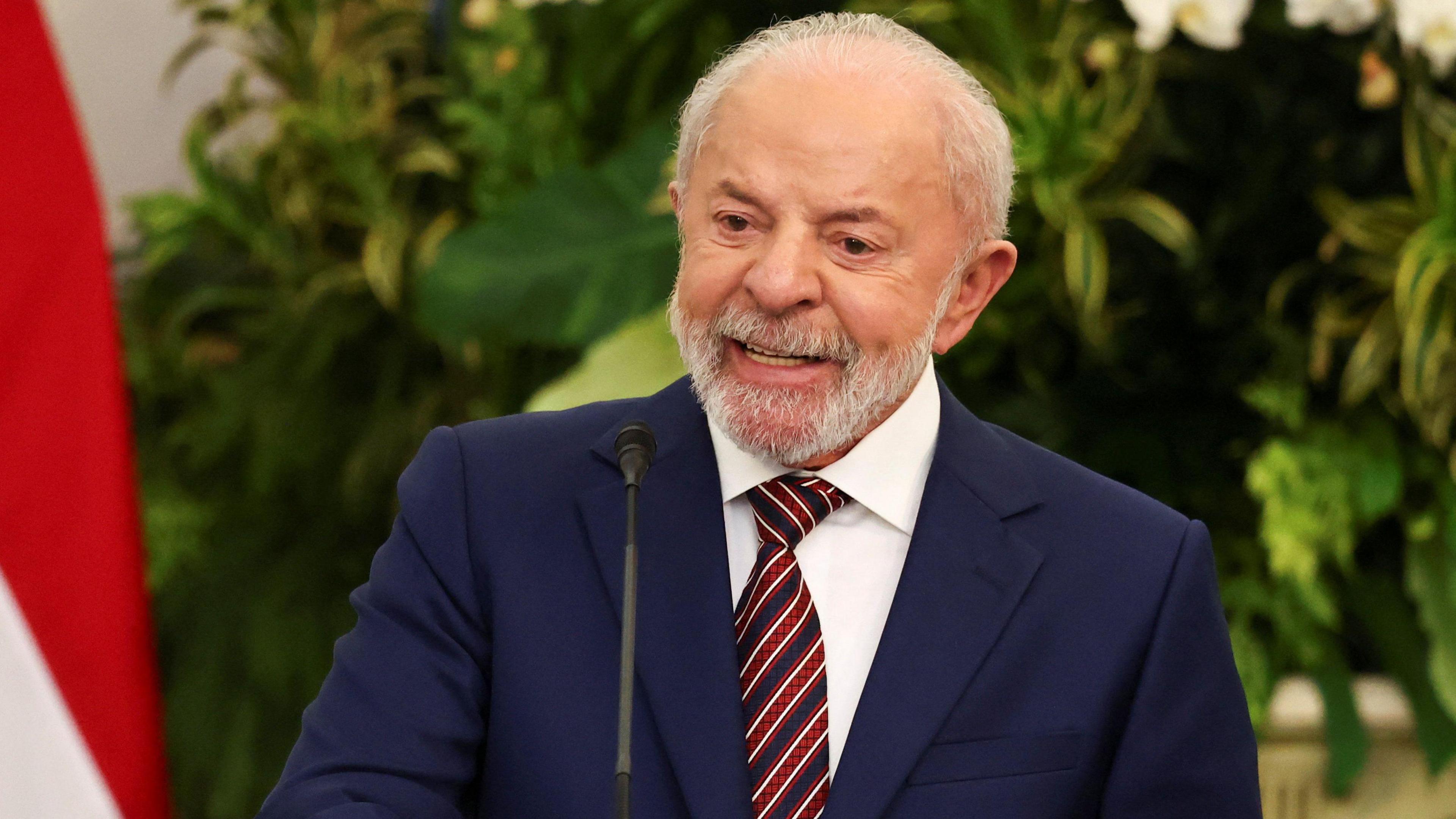 Lula is wearing a fitted navy suit with a white shirt on underneath. He is wearing a funky red, white and blue striped tie and is standing behind a lectern in mid speech. He has a grey beard and grey hair.