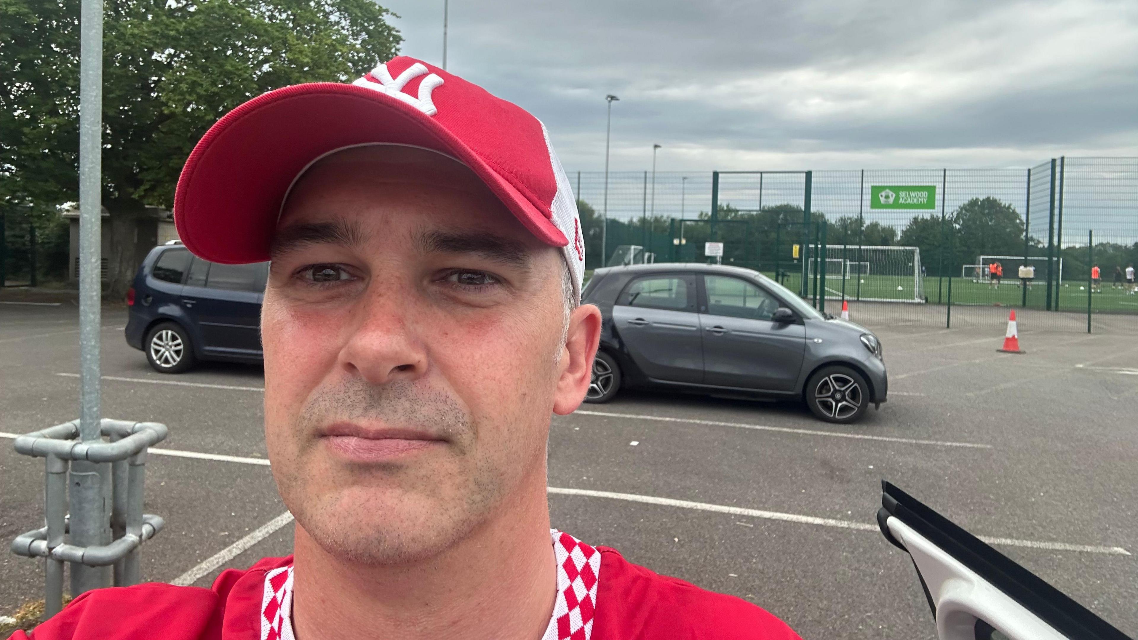 Man in red baseball hat. He's stood in a car park by a football playing field. It's a selfie and he's got a red top on.