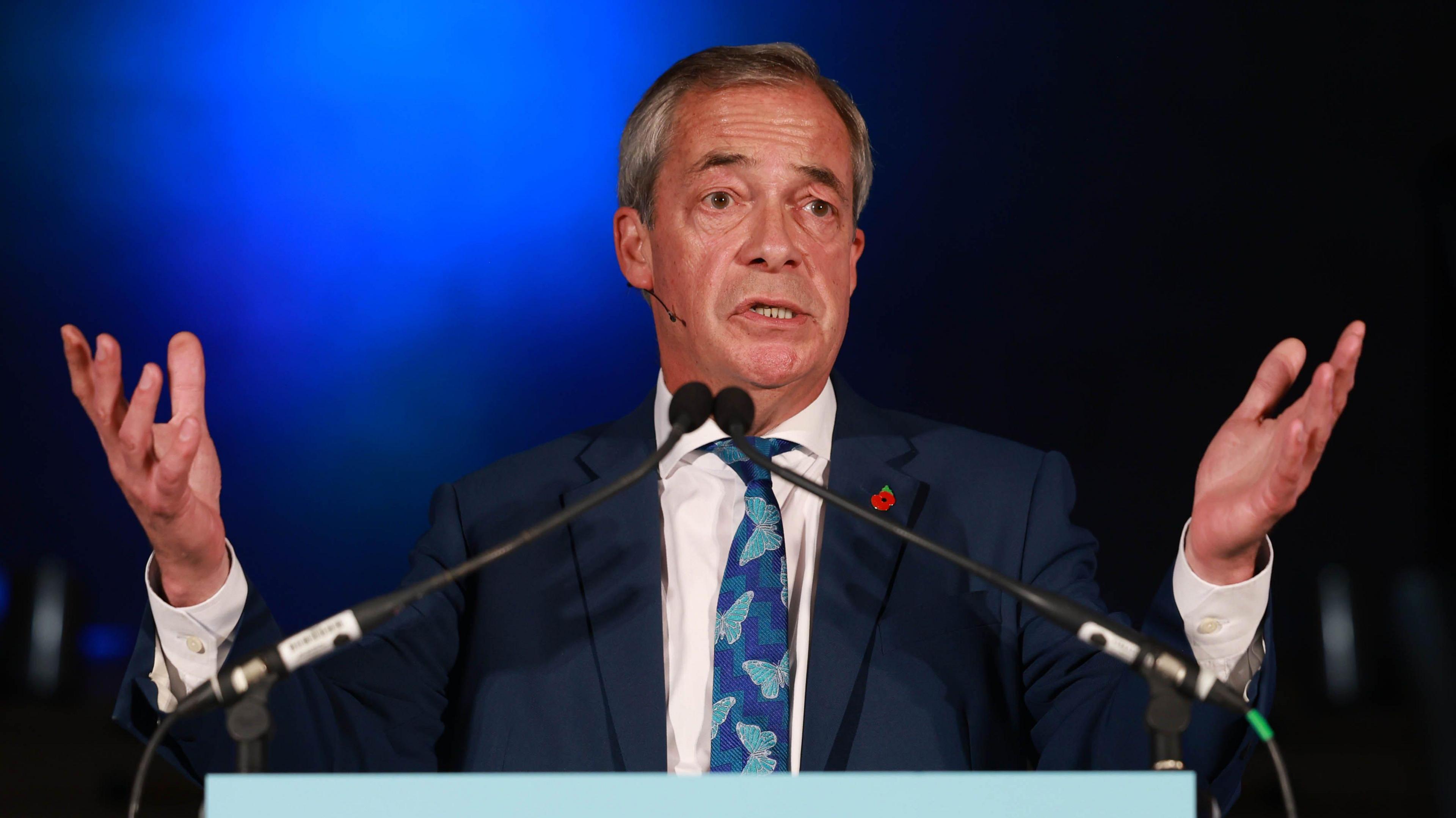 Nigel Farage stands on a stage and speaks into two microphones. He has both hands raised in front of him as he talks and looks to the right. He wears a blue suit, white shirt and blue tie with butterflies on it. 
