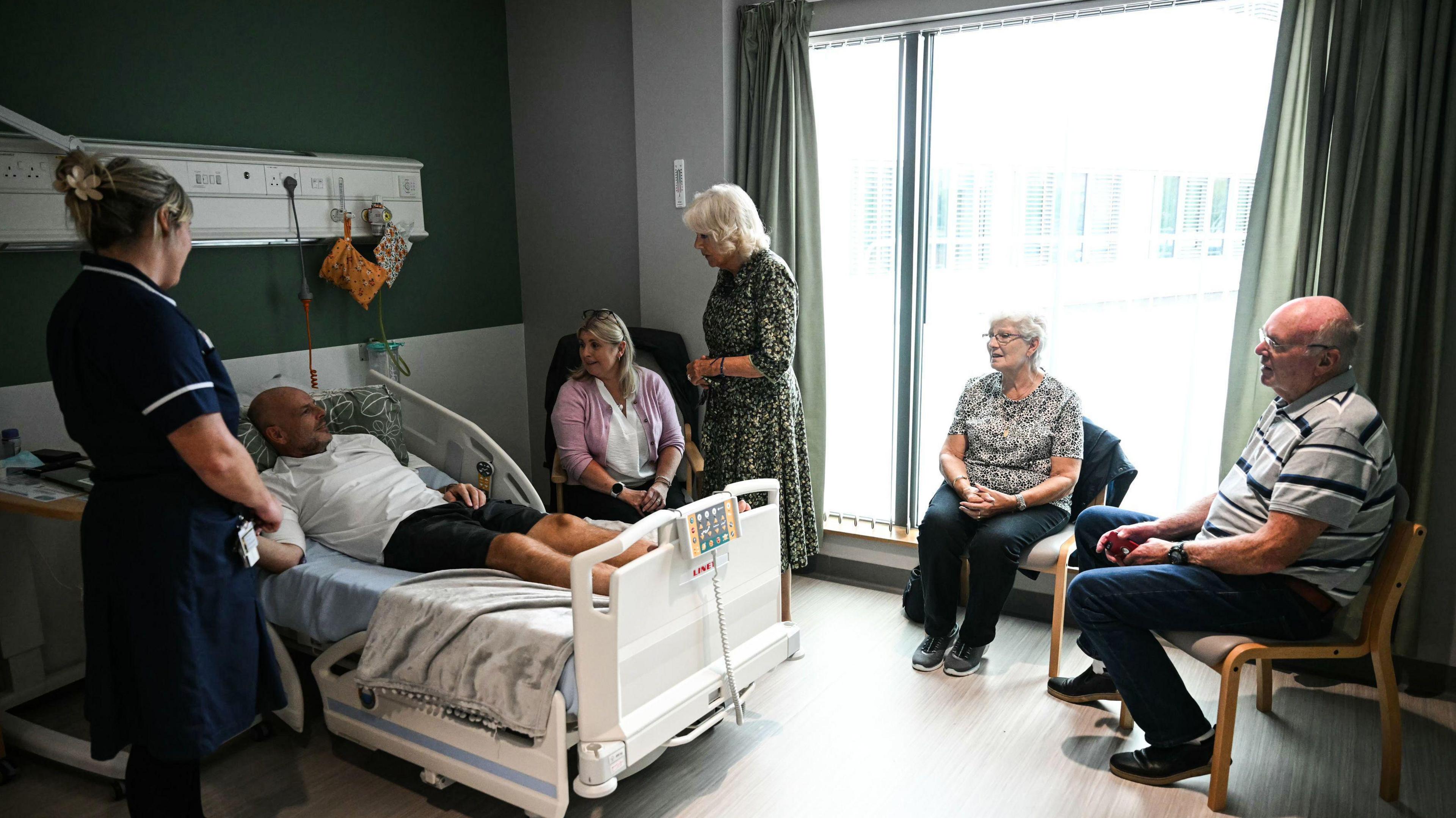 Queen Camilla stands next to hospital bed speaking to Paul Holdway, who has family members around him