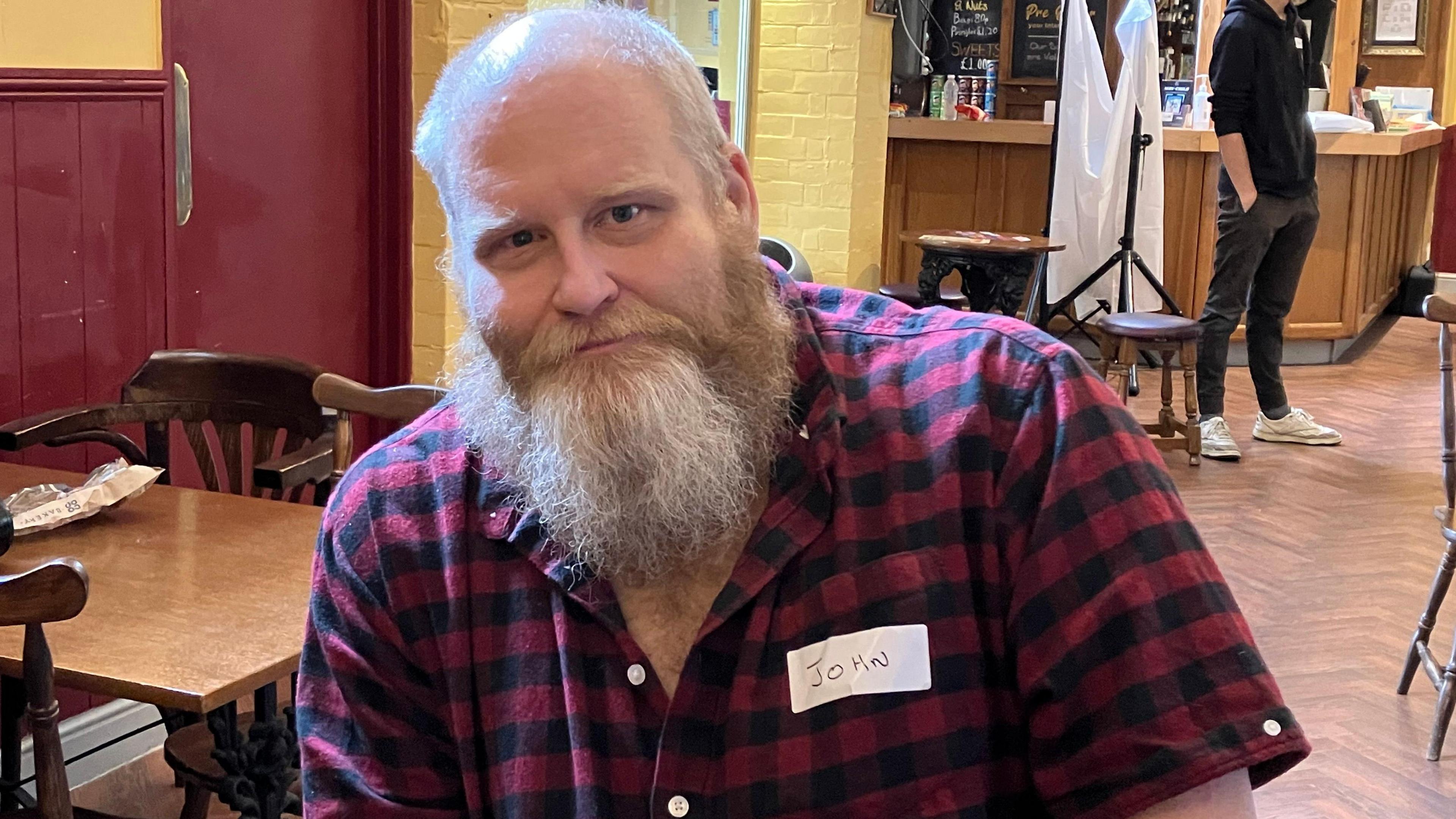 Joh Hales smiles and looks directly at the camera, he has a beard and is wearing a checked red shirt and sitting a cafe with a kiosk behind him