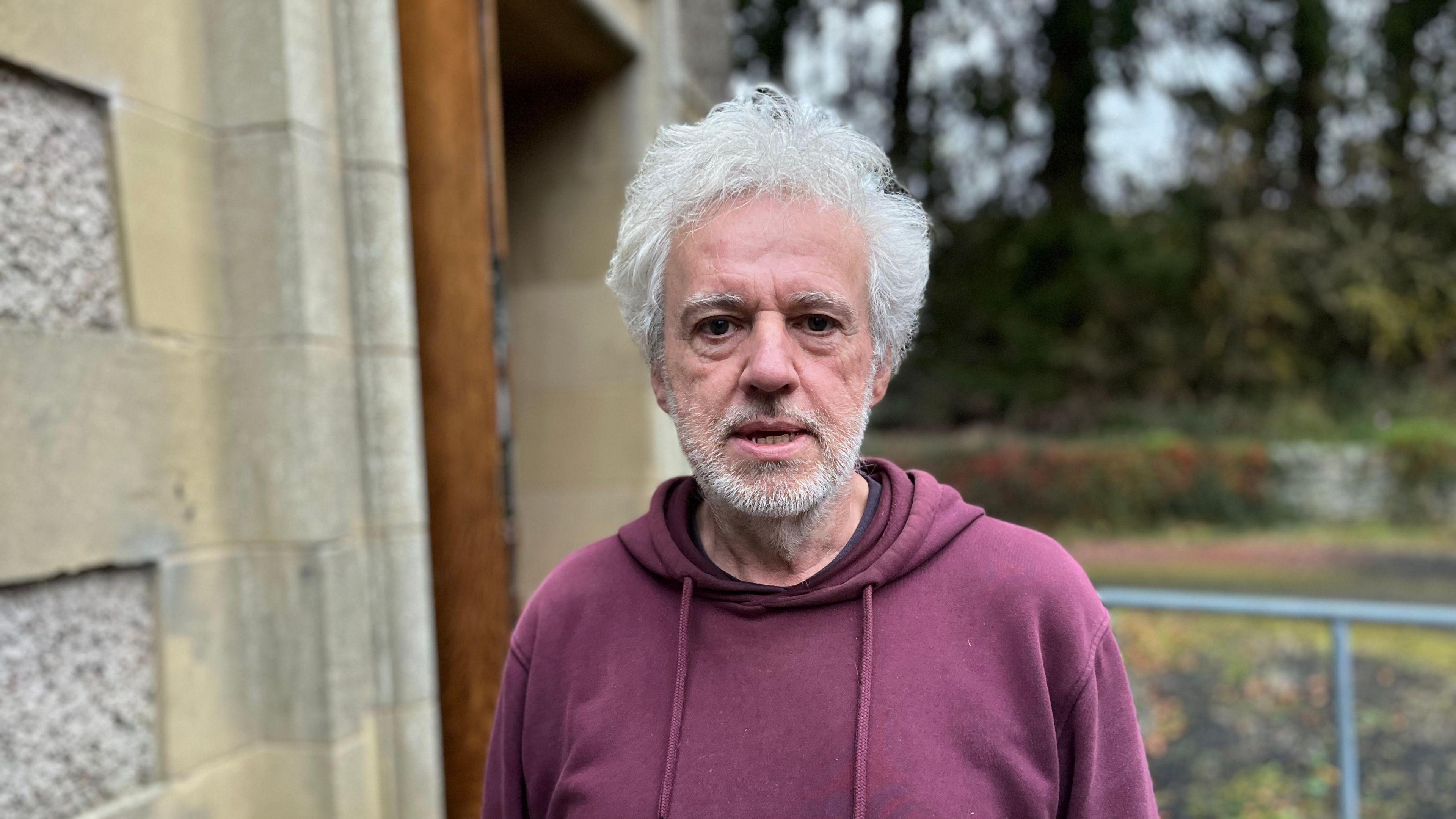 A grey-haired man with a wine-coloured hoodie standing outside a village hall. There are trees and a metal railing behind him.