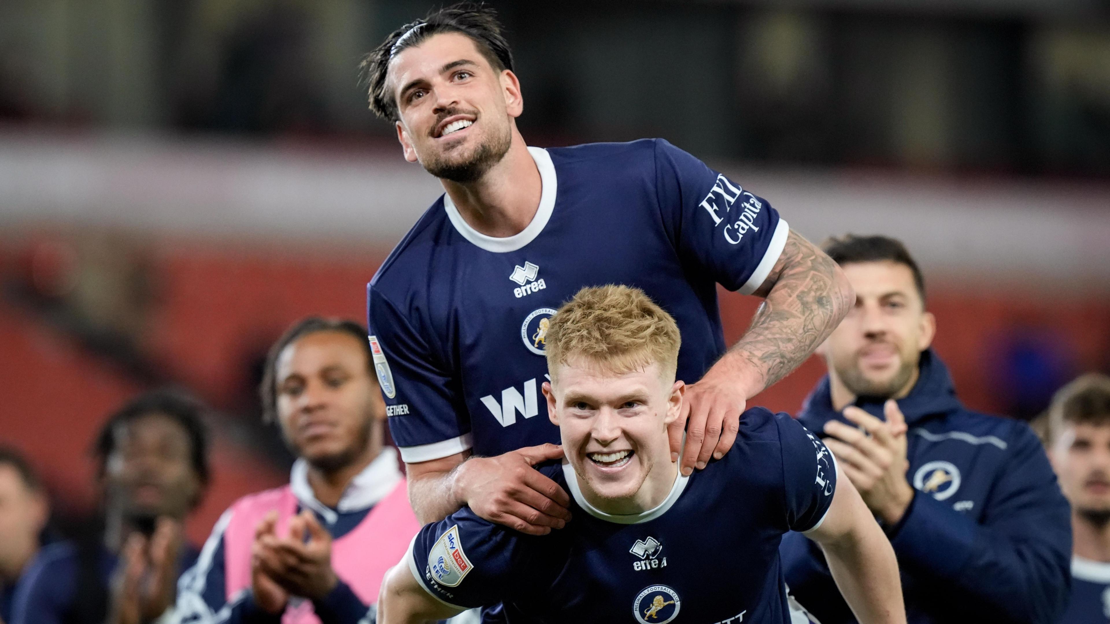 Tristan Crama climbs on Josh Coburn's back, with both wearing Millwall's navy blue home shirt and smiling after the win at Stoke