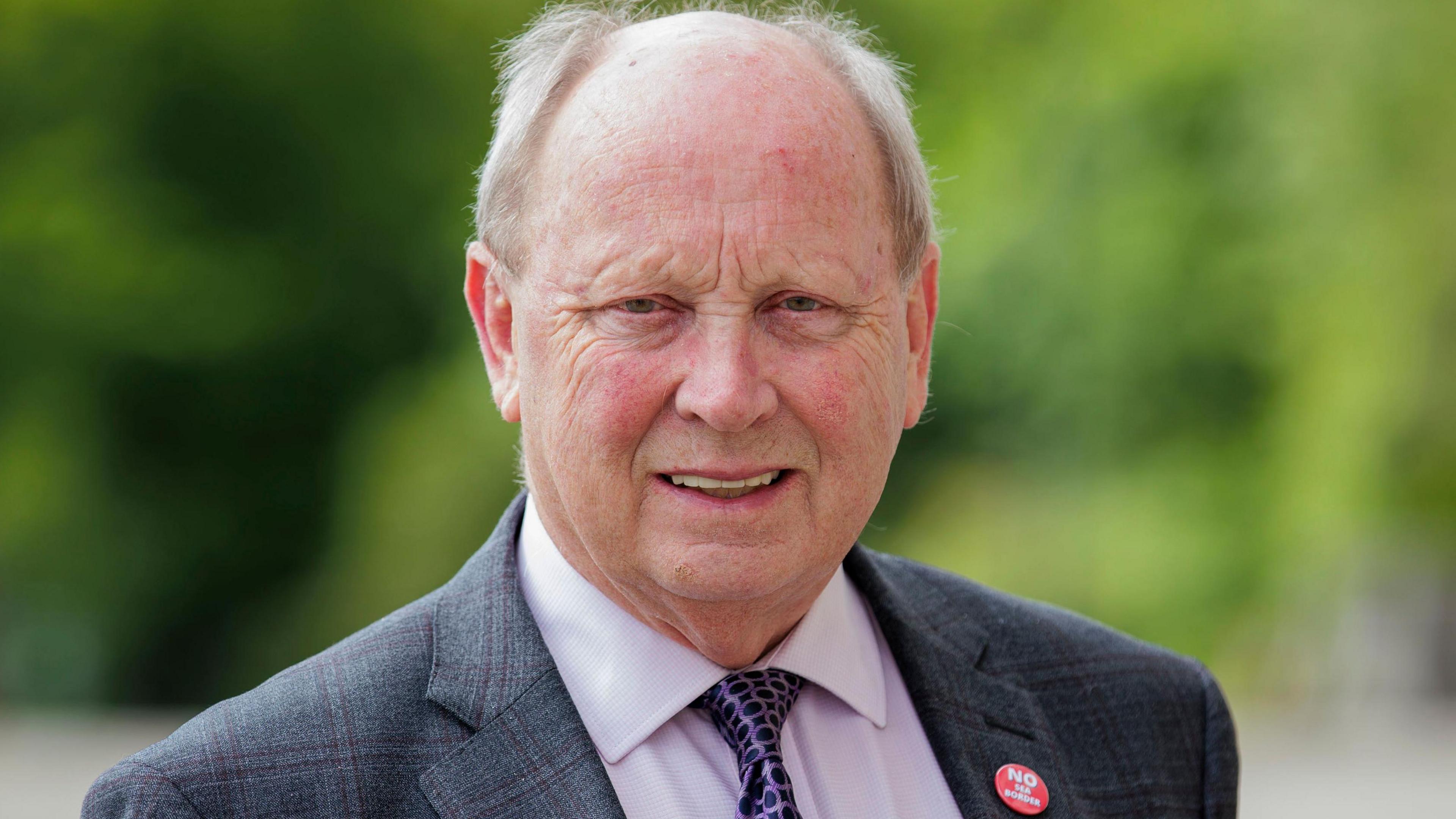 Jim Allister in a grey checked blazer smiles to camera. He has grey hair and is wearing a lilac shirt with purple spotted tie. On his right lapel he wears a badge reading 'no sea border'. 