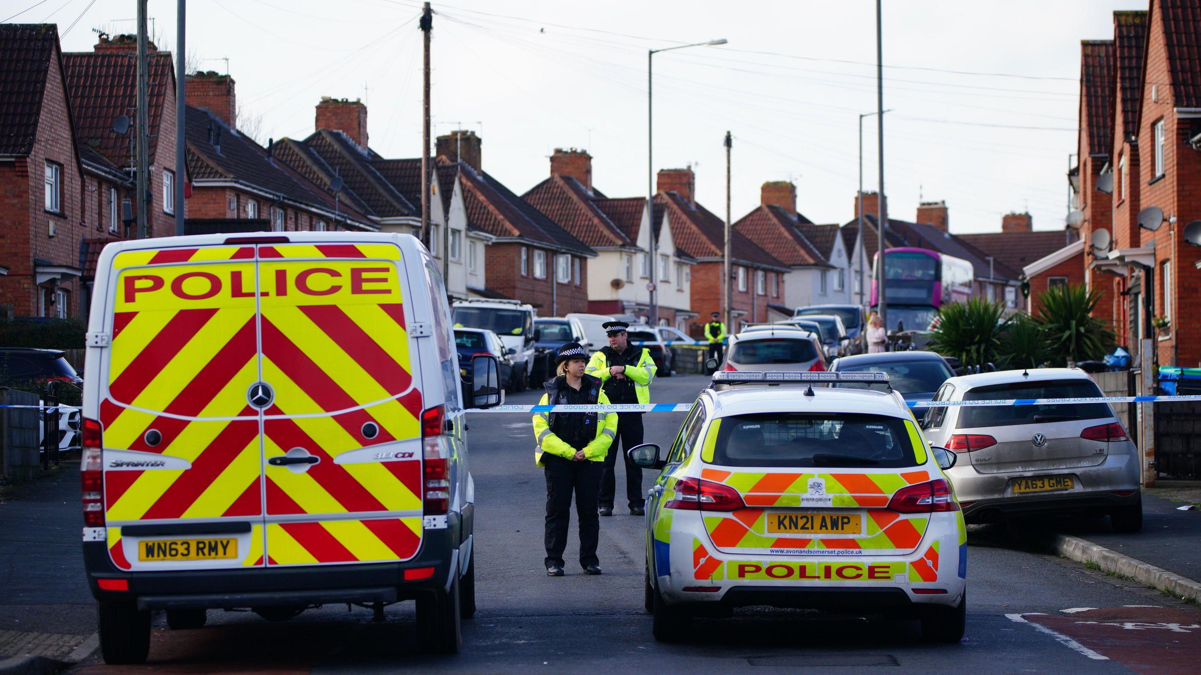 A group of police vehicles parked on a street in Knowle West in Bristol. A couple of officers are visible wearing high-viz jackets and there is blue and white police tape stretched across the street