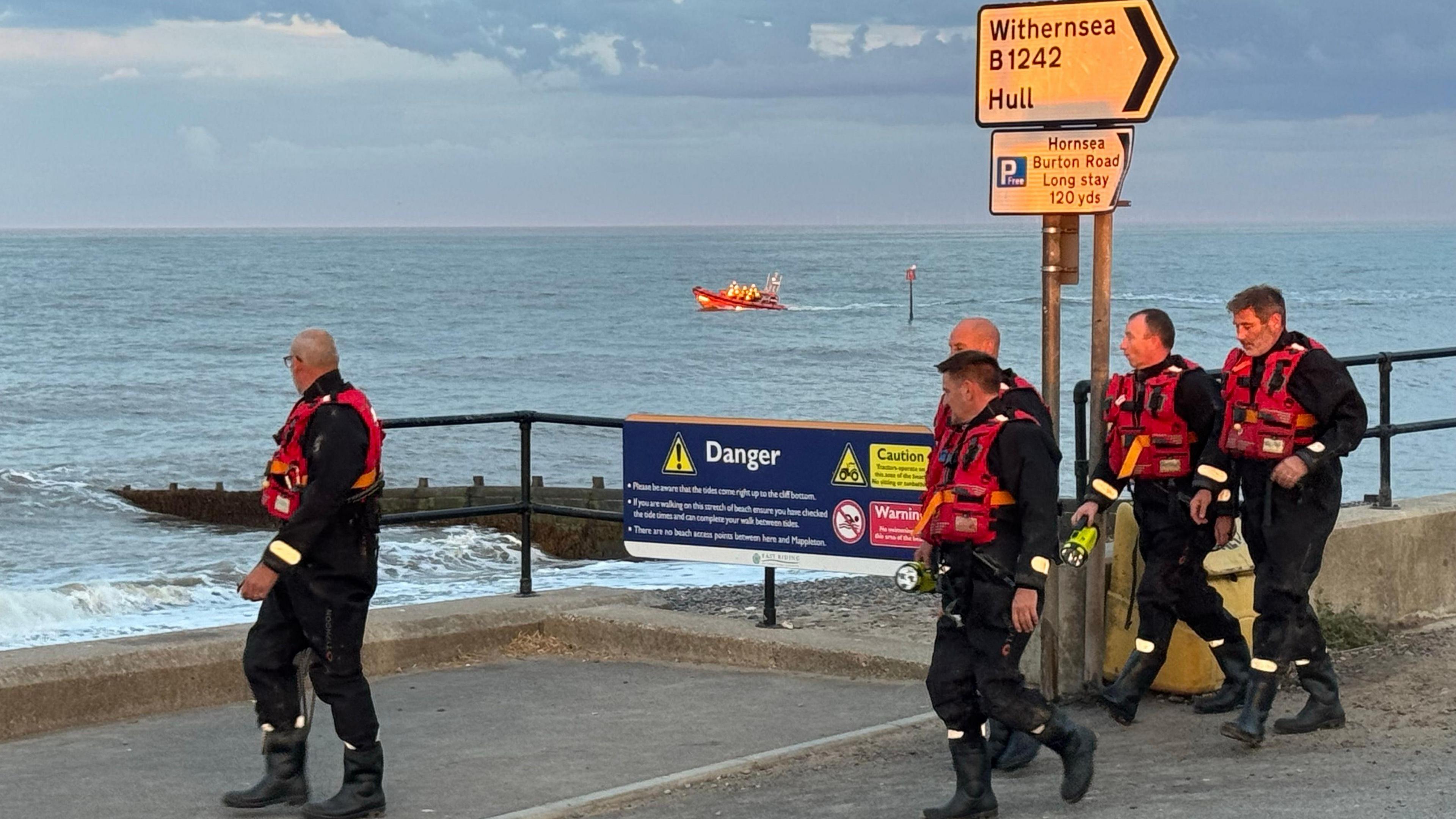 Rescue crew in protective equipment walking along the seafront. There is a lifeboat visible on the sea in the distance.