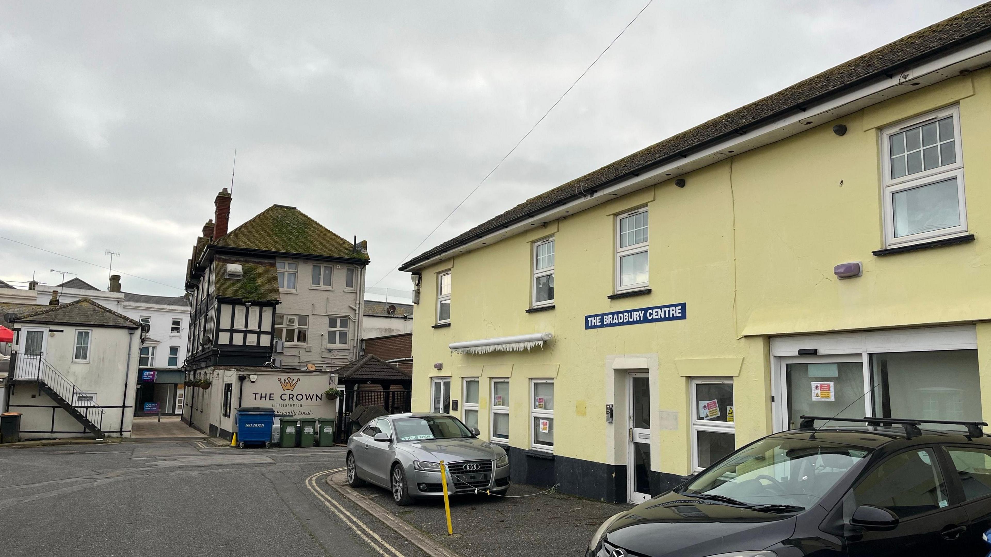 A yellow building where the mosque is proposed to open in. It has a small number of parking spaces which have cars parked in them. In the background is a pub called The Crown and an alleyway leading to the main shopping high street.