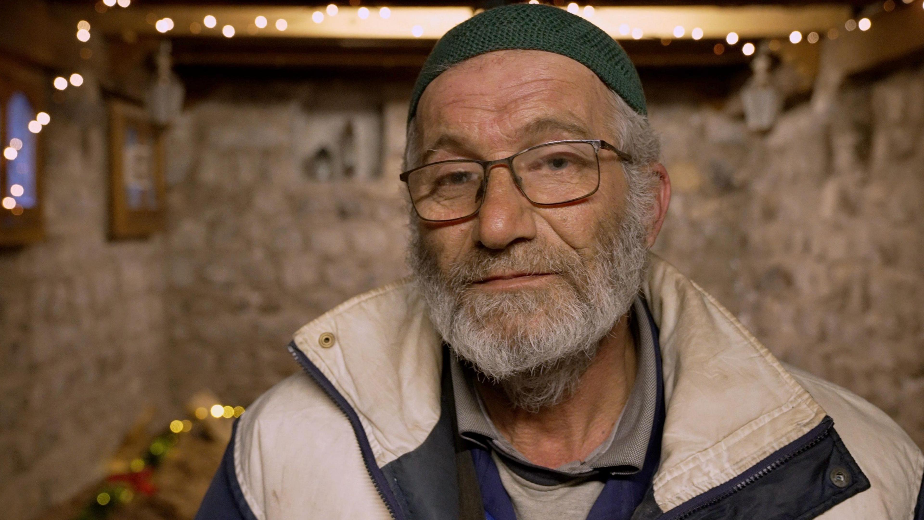 Farhan Abdul Ghani stands in front of camera wearing a hat and glasses, and a white beard, inside a stone-walled room