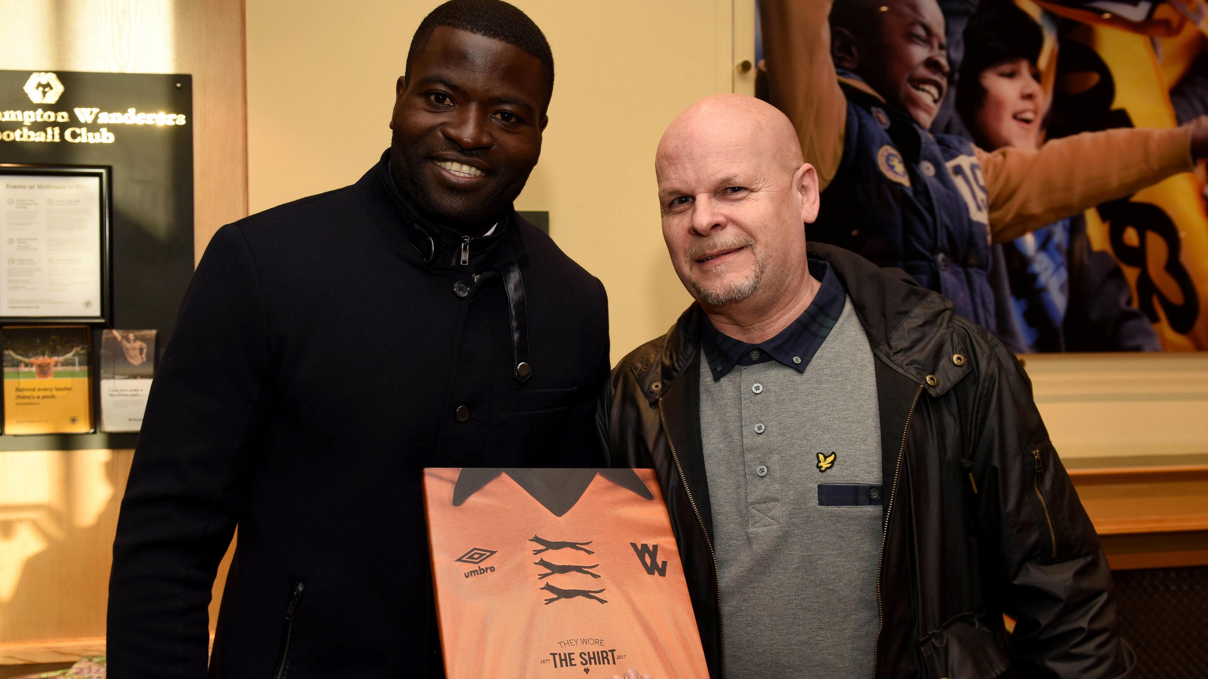 Two men in the reception of Wolves football club. Both wearing coats. One has a copy of a book about football shirts