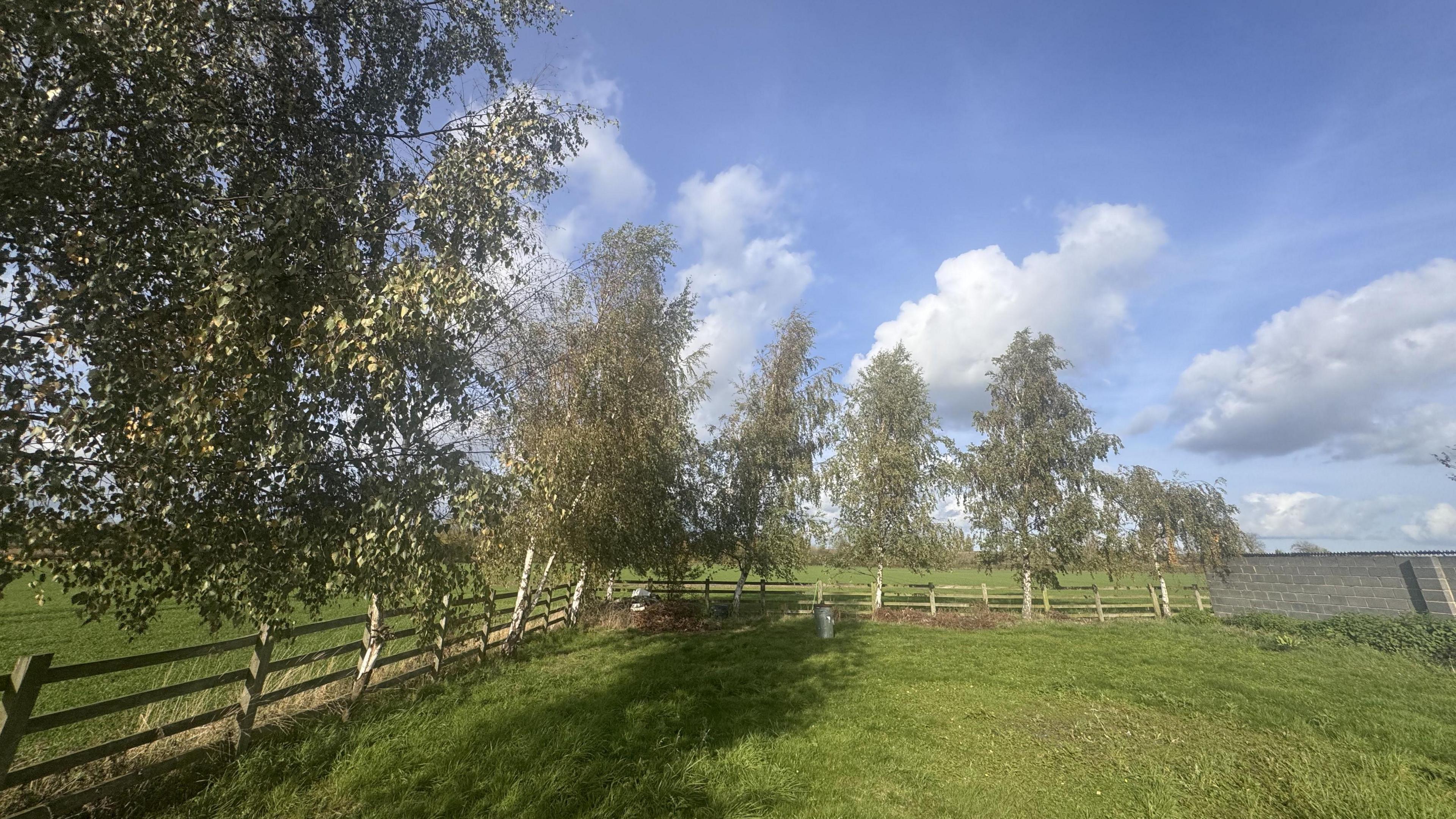 The pictures is of a fenced off field, with further fields behind it. There are trees grown around the fence, and there's a grey brick structure to the right of the image. It's a sunny day, and the sky is blue with scattered clouds.
