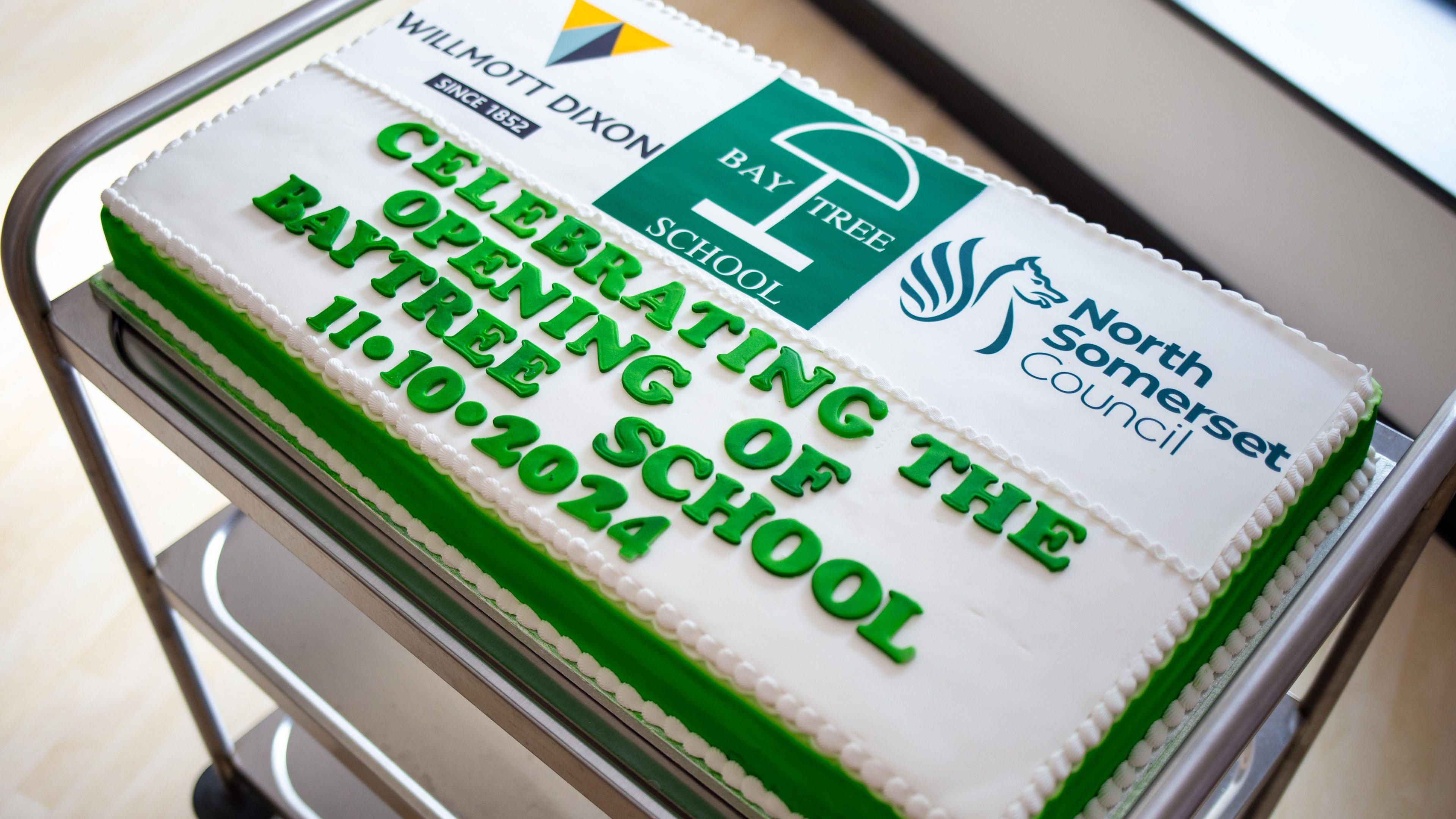 A picture of a white cake on a trolley which reads: "Celebrating the opening of Baytree School. 11.10.2024" in green lettering