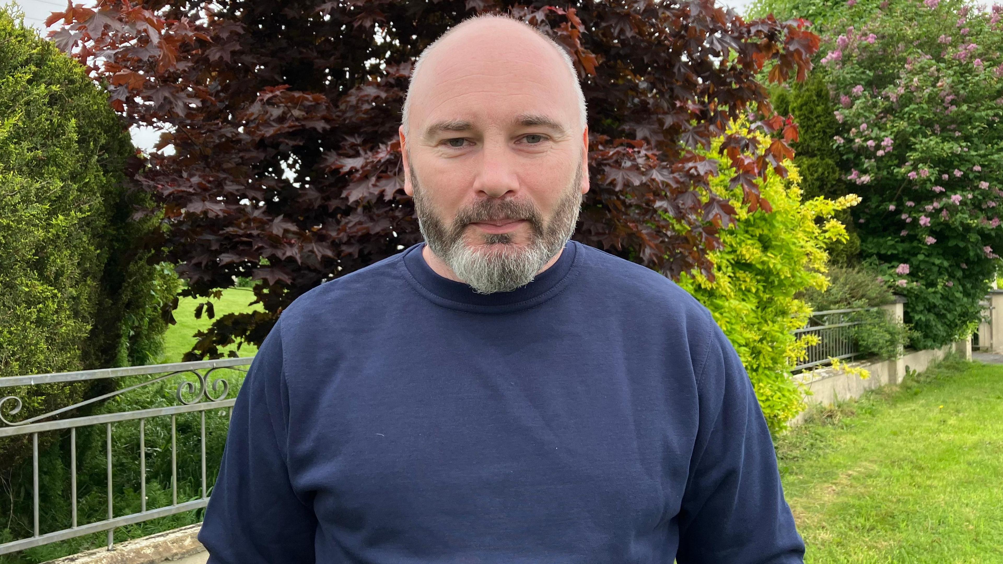 Lee Williamson stands in his garden. he is wearing a blue sweater, railings and trees can be seen in the background