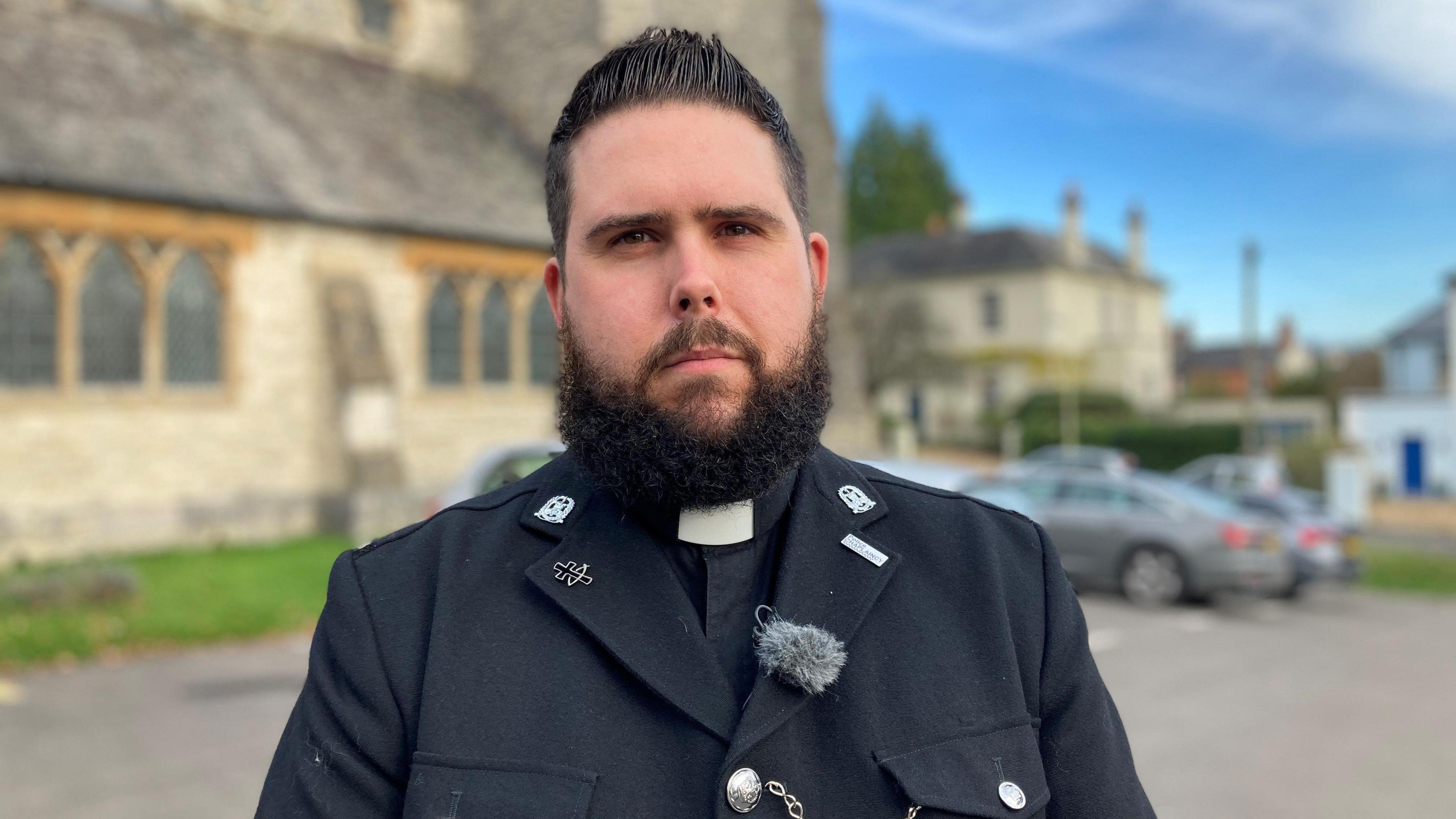 A bearded man wearing a white clerical collar and a black jacket, looking directly at the camera. There's a church and a car park in soft focus behind him.