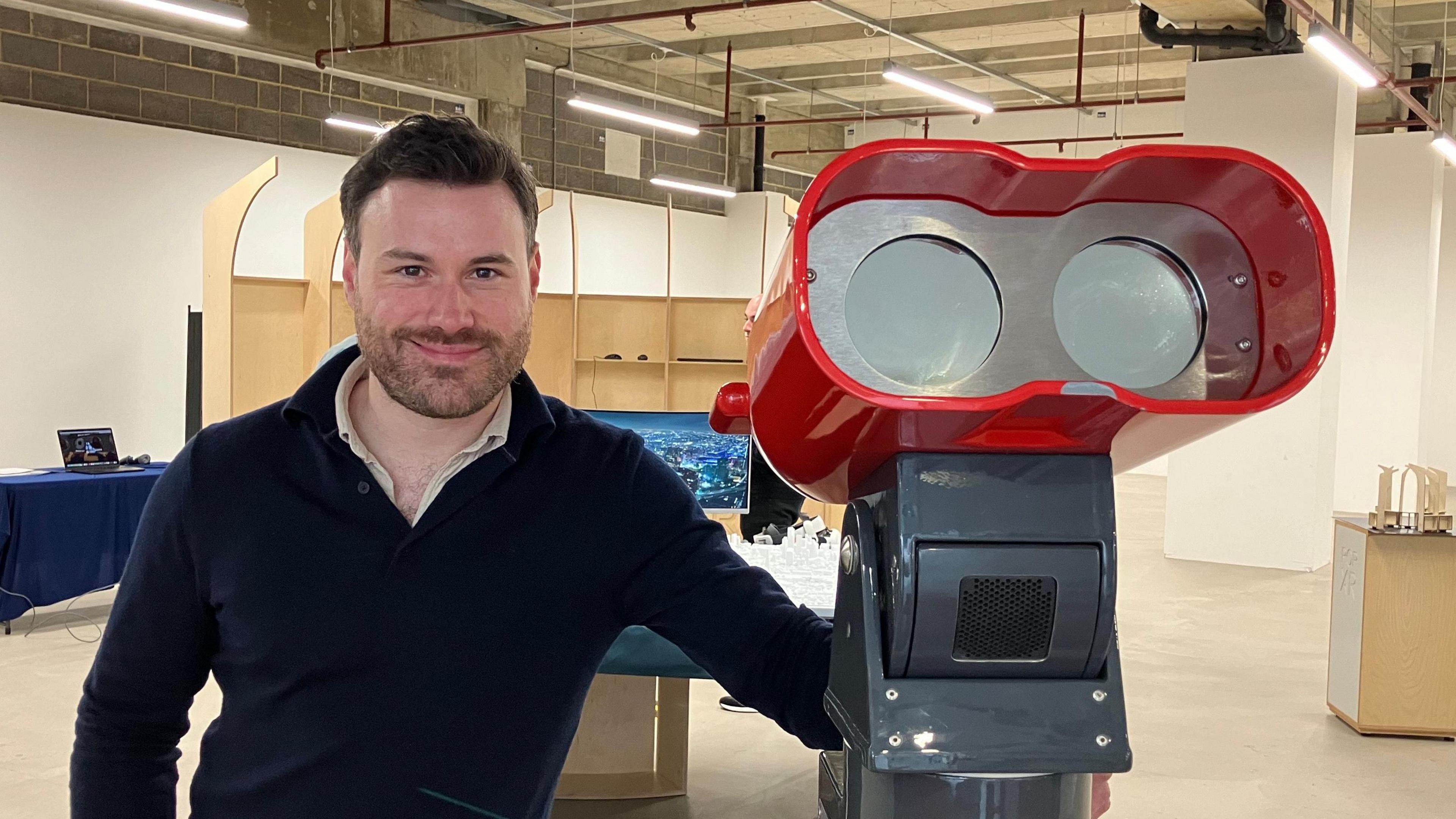 Jack Norris smiles at the camera while standing next to a large part of red binoculars on a plinth. He has short dark hair and wears a black jumper with a white shirt underneath.