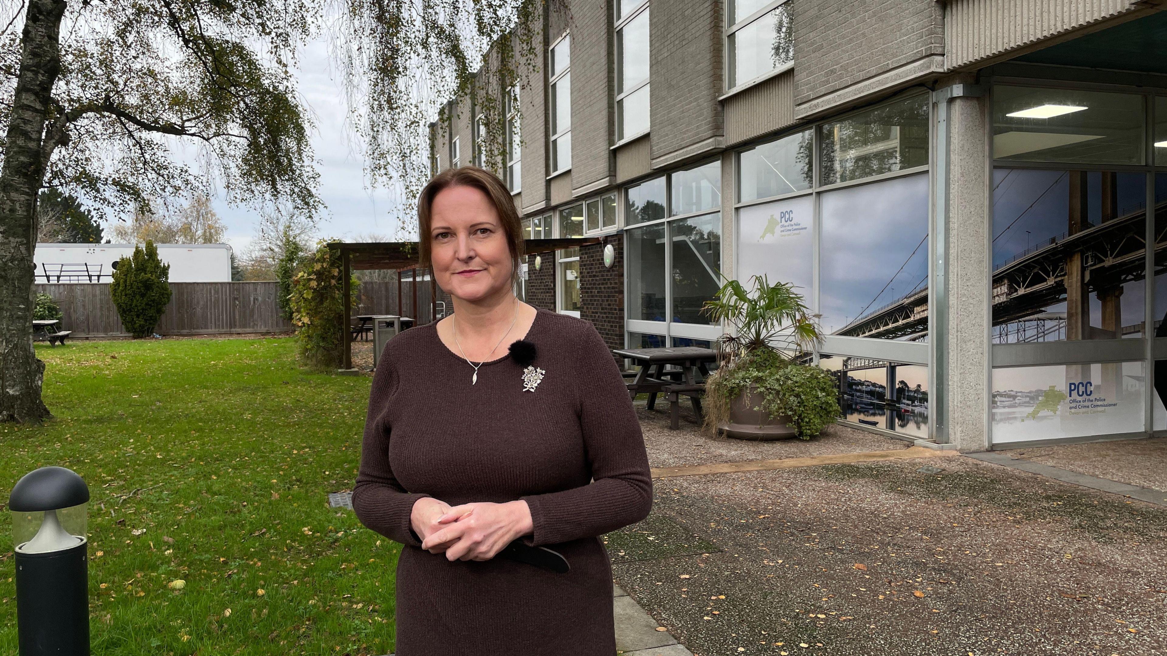 Alison Hernandez stands outdoors on a path next to a patch of grass. She is holding her hands together and is smiling a the camera. She is wearing a brown long-sleeved dress with a brooch. A tree is on the grass. A grey building with large windows is behind her.