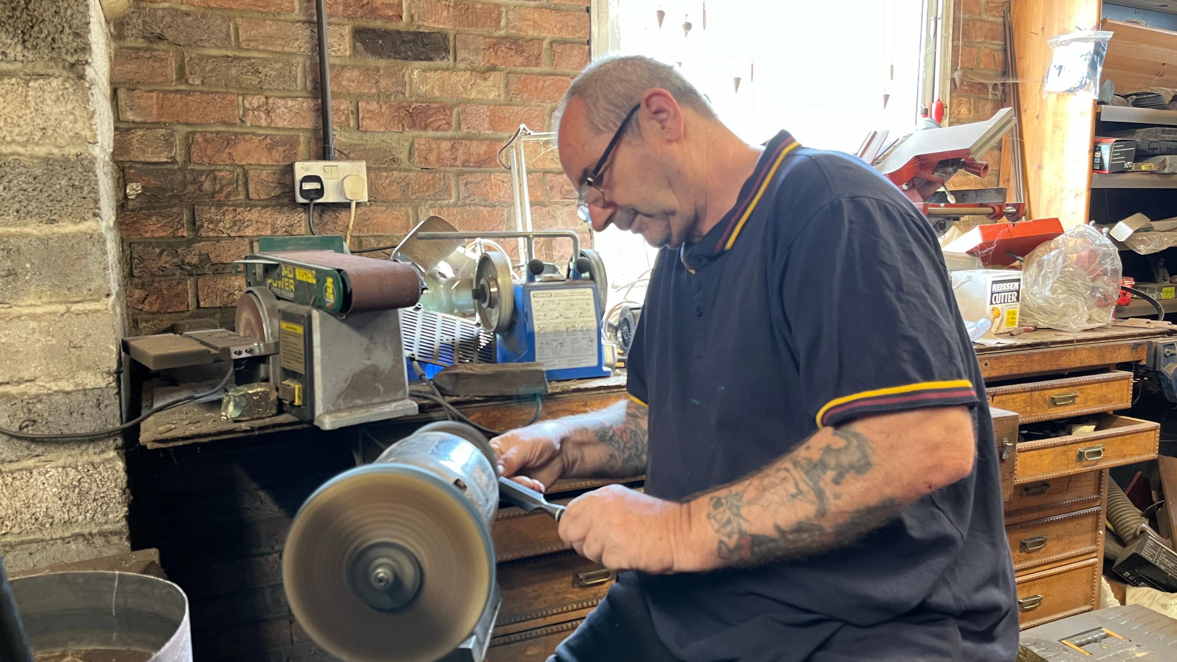 David Nicholas wearing a dark blue T-shirt which has a red and yellow stripe on the collar and sleeves. He is in his workshop sitting next to a piece of machinery. He is looking away from the camera and has protective eyewear on.