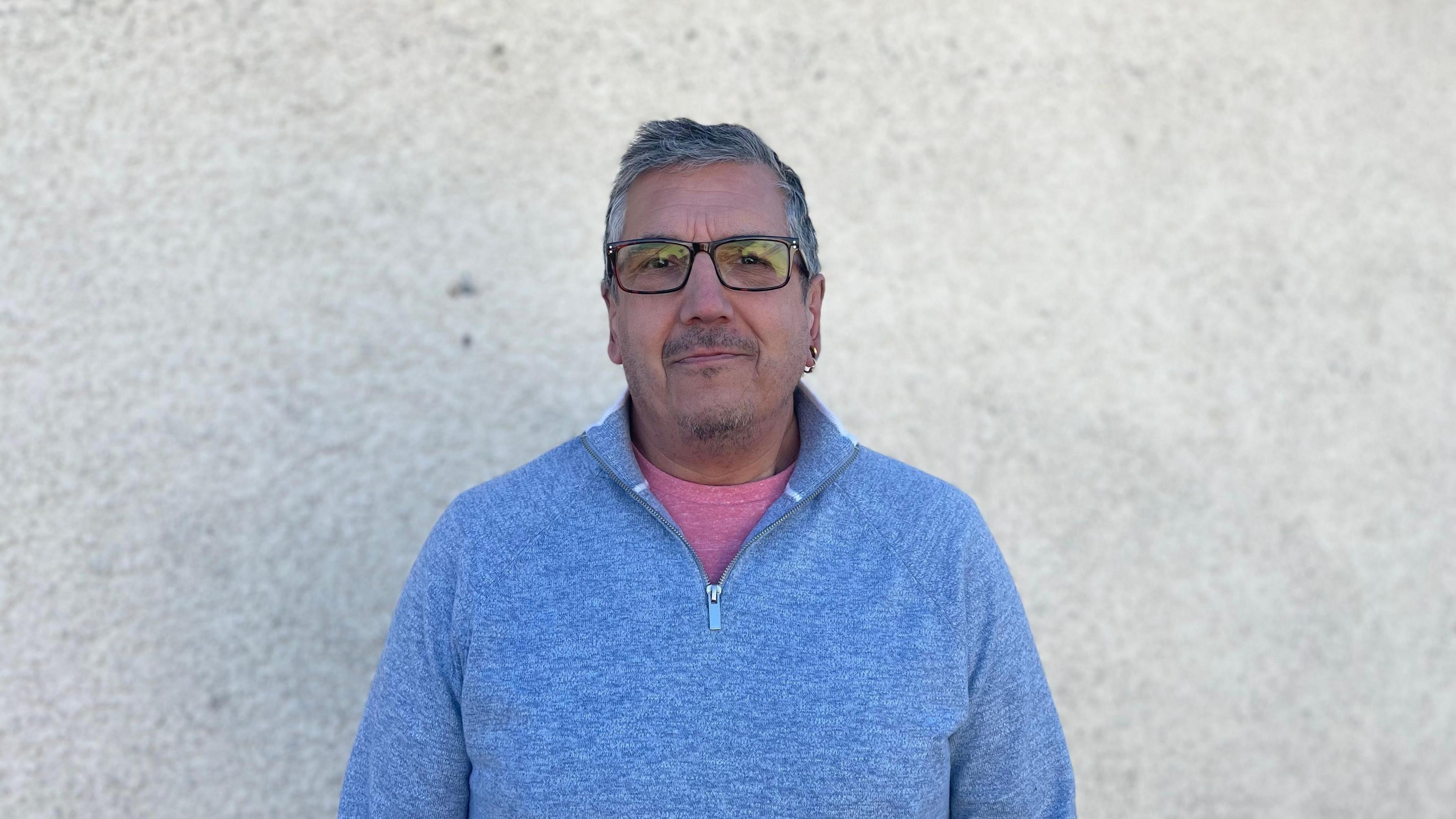 A man in a blue quarter zip stand in front of a white wall. He has a pink T-shirt underneath. He wears black glasses and two gold earrings in his left ear. 