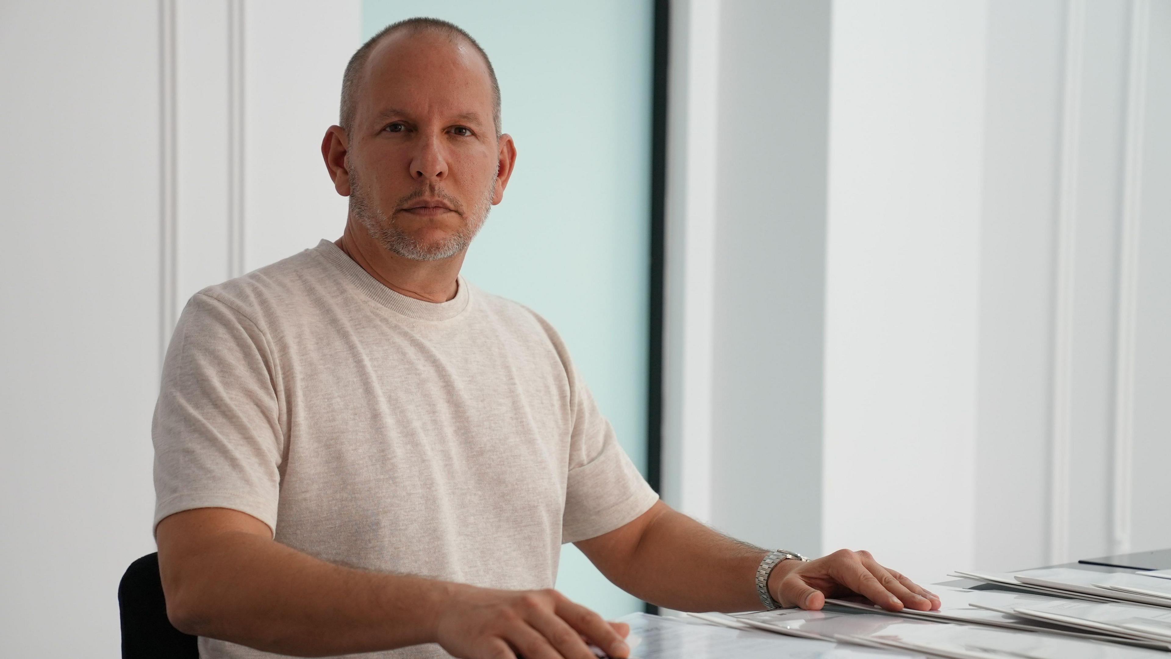A man sits at a desk wearing a light T-shirt with his hands on the table