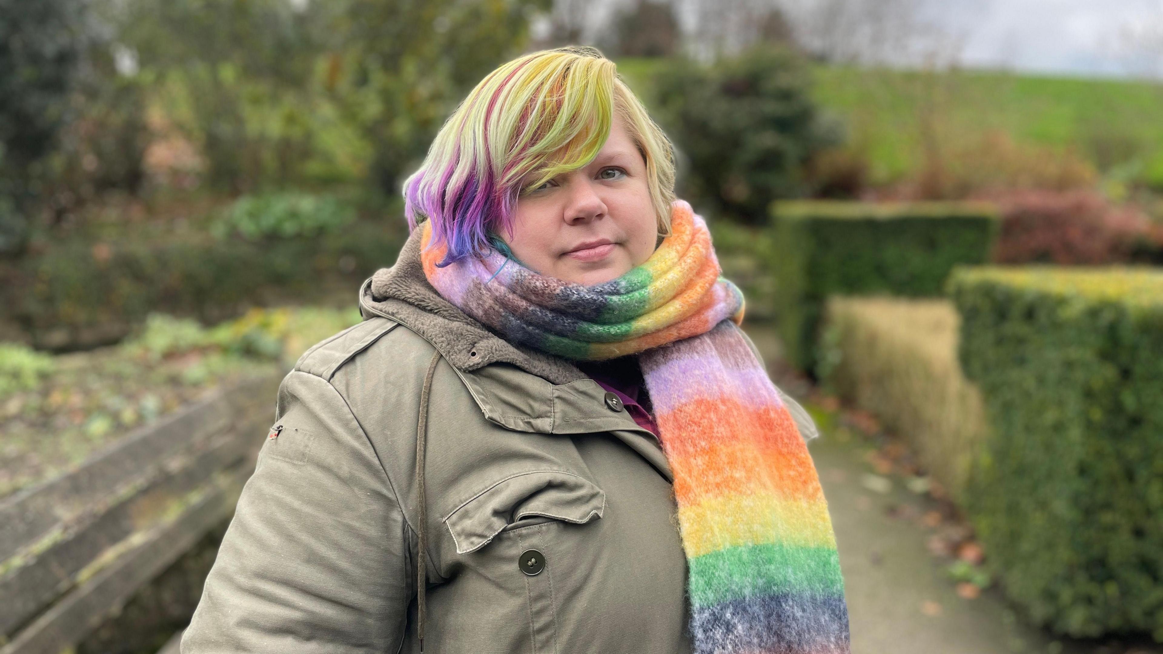 Rachel (a woman with multi-coloured hair, a rainbow scarf and a brown coat) looks directly into the camera