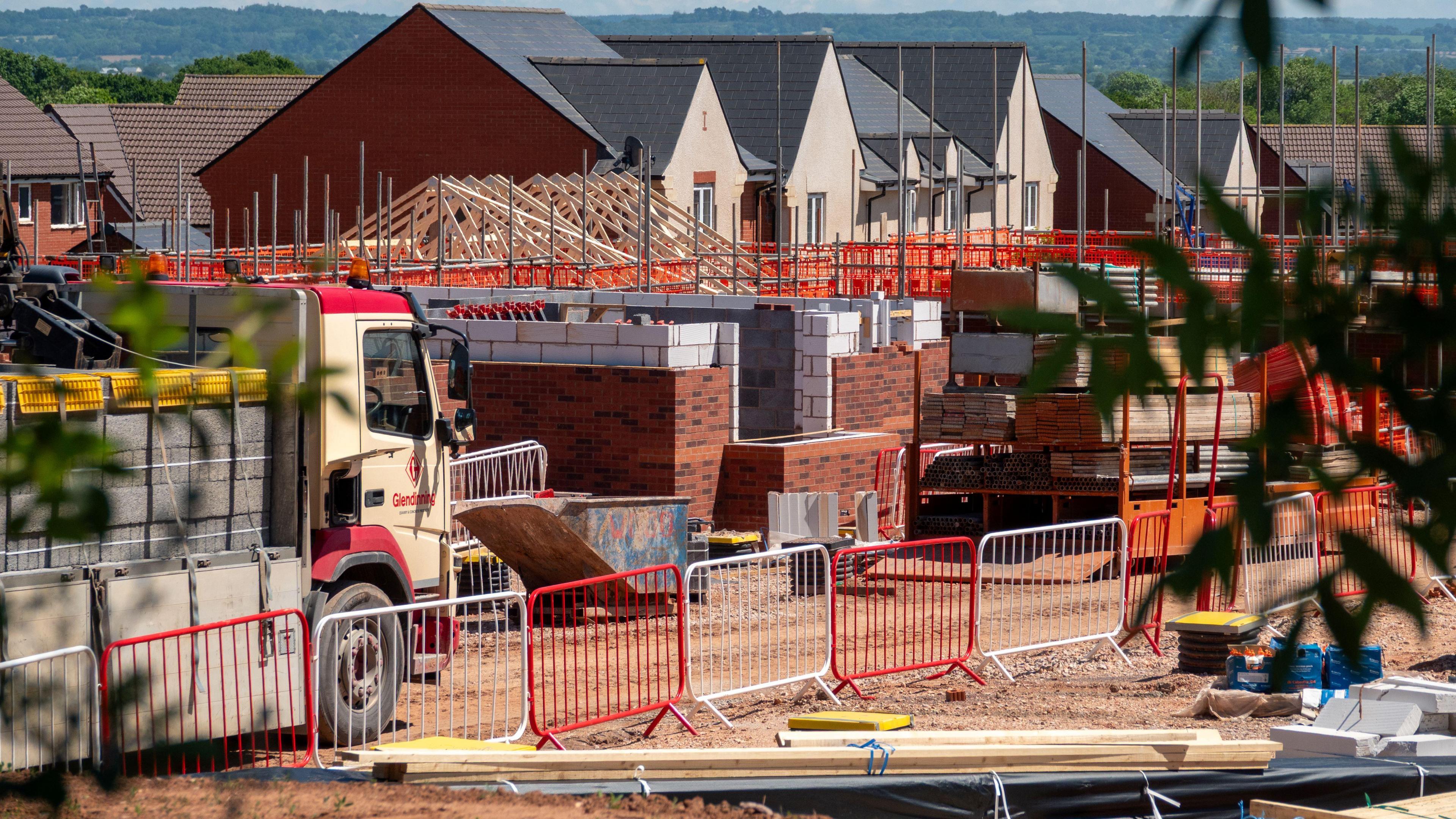 A housing construction site with a number of partially built houses. There is a lorry in the foreground carrying construction materials, and fencing surrounds the site.