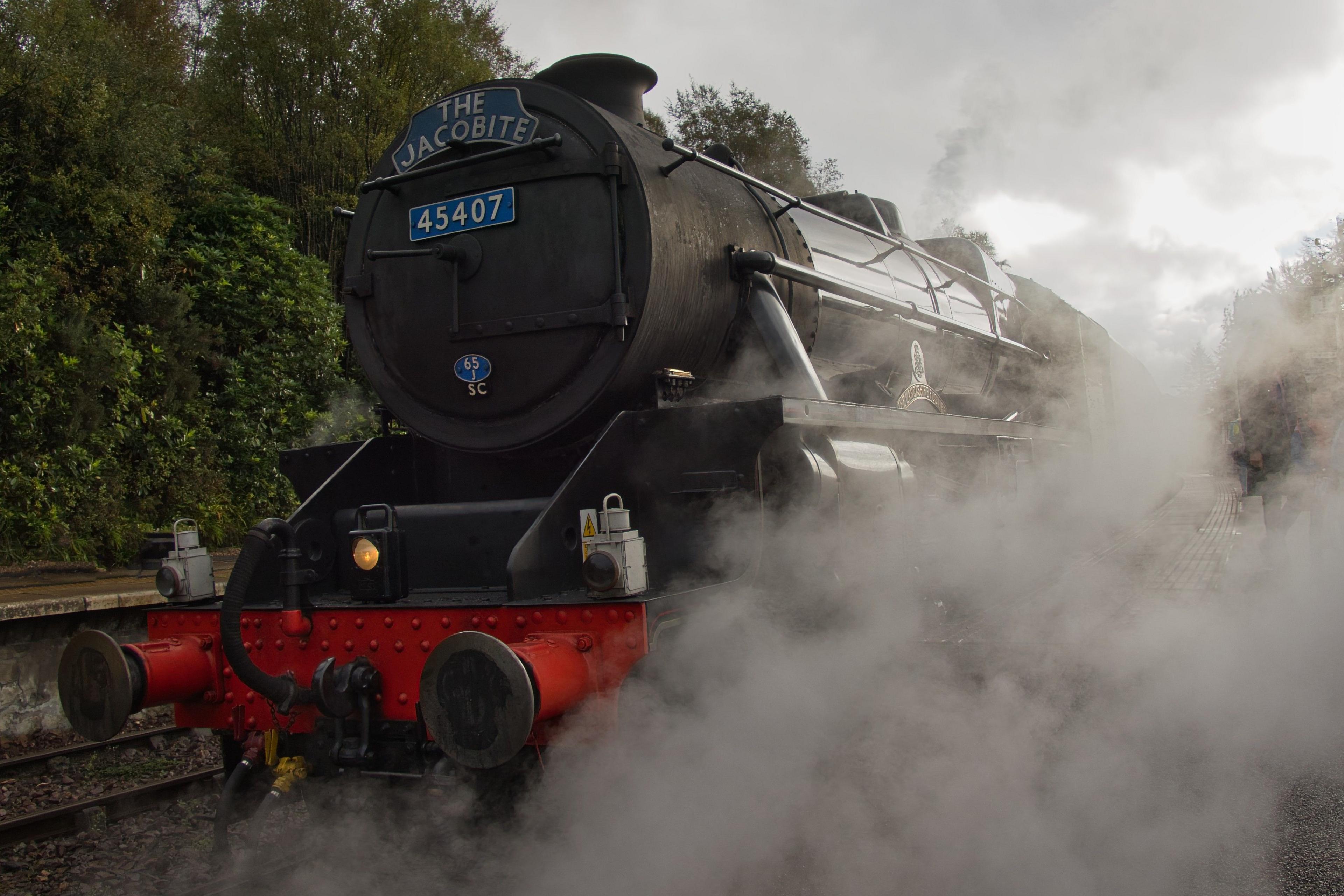 The black steam locomotive "The Jacobite” stands on a railway track, surrounded by thick clouds of white steam. Its front features bright red buffers and metallic details, contrasting with the dark body of the train. Dense greenery lines the platform in the background.