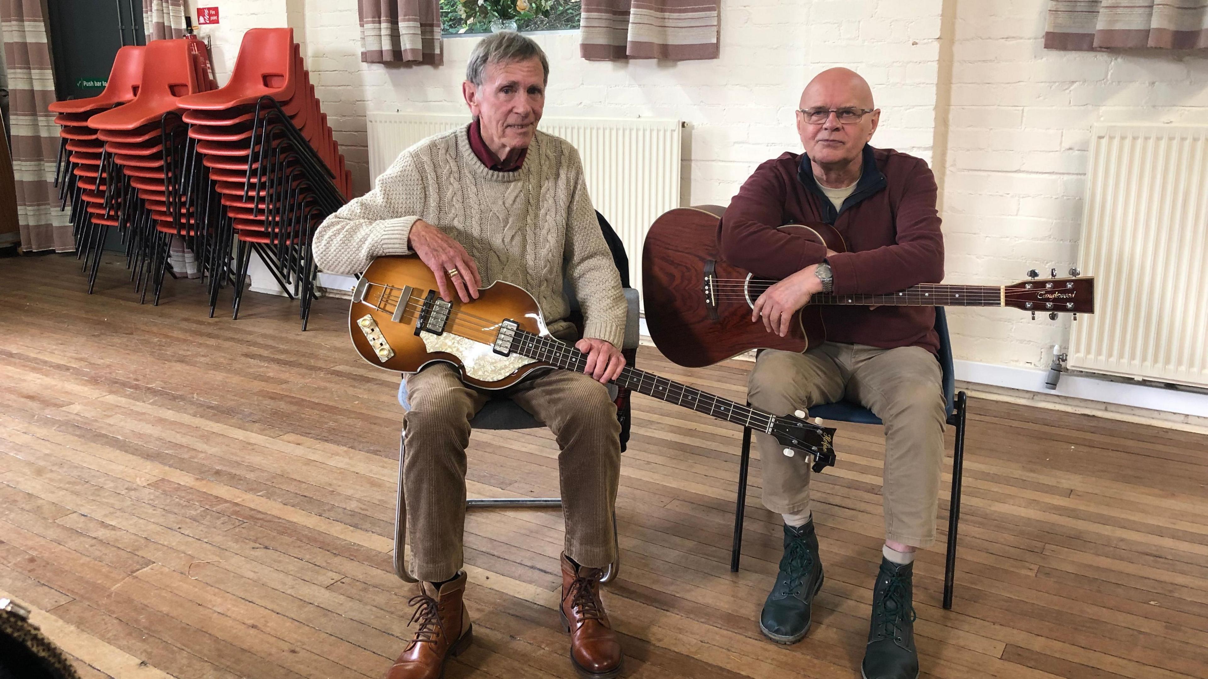 Two men sitting side by side on blue plastic chairs. The man on the left is wearing a white jumper and light brown trousers. He is holding an electric bass guitar. The man on the right is wearing a maroon jumper and light brown trousers. He is holding a brown  semi acoustic six-string folk guitar. The floor is made out of wood. Behind them to the left are three stacks of red plastic chairs. The wall behind them is cream and made of brick. 