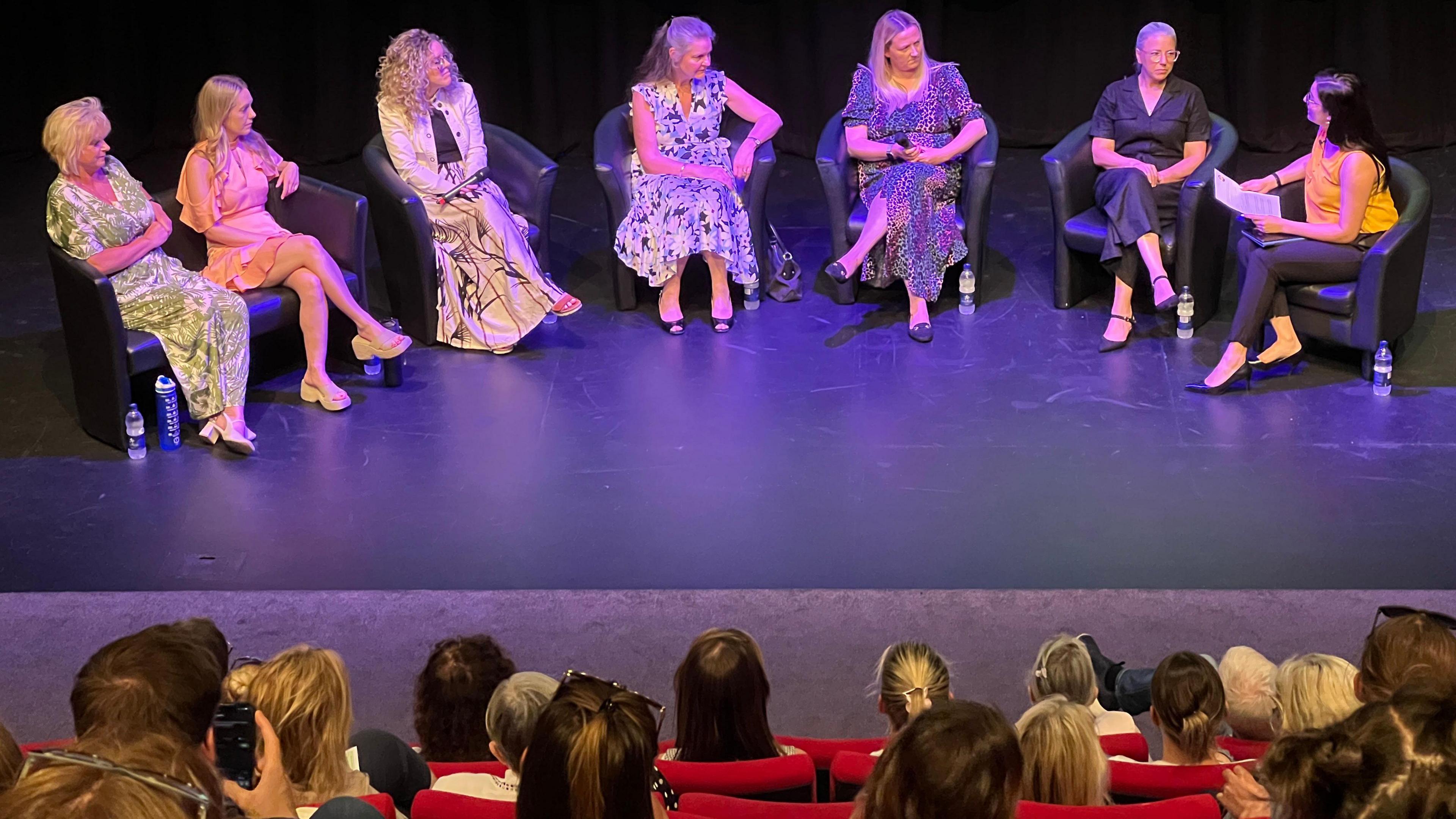 A panel discussion is taking place on a stage with seven individuals seated in a row of black chairs. The individuals are dressed in various outfits, including dresses. There are water bottles placed on the floor next to some of the chairs. The audience is seated in red theatre seats, facing the stage. The background features a large screen with a gradient colour display at the top.