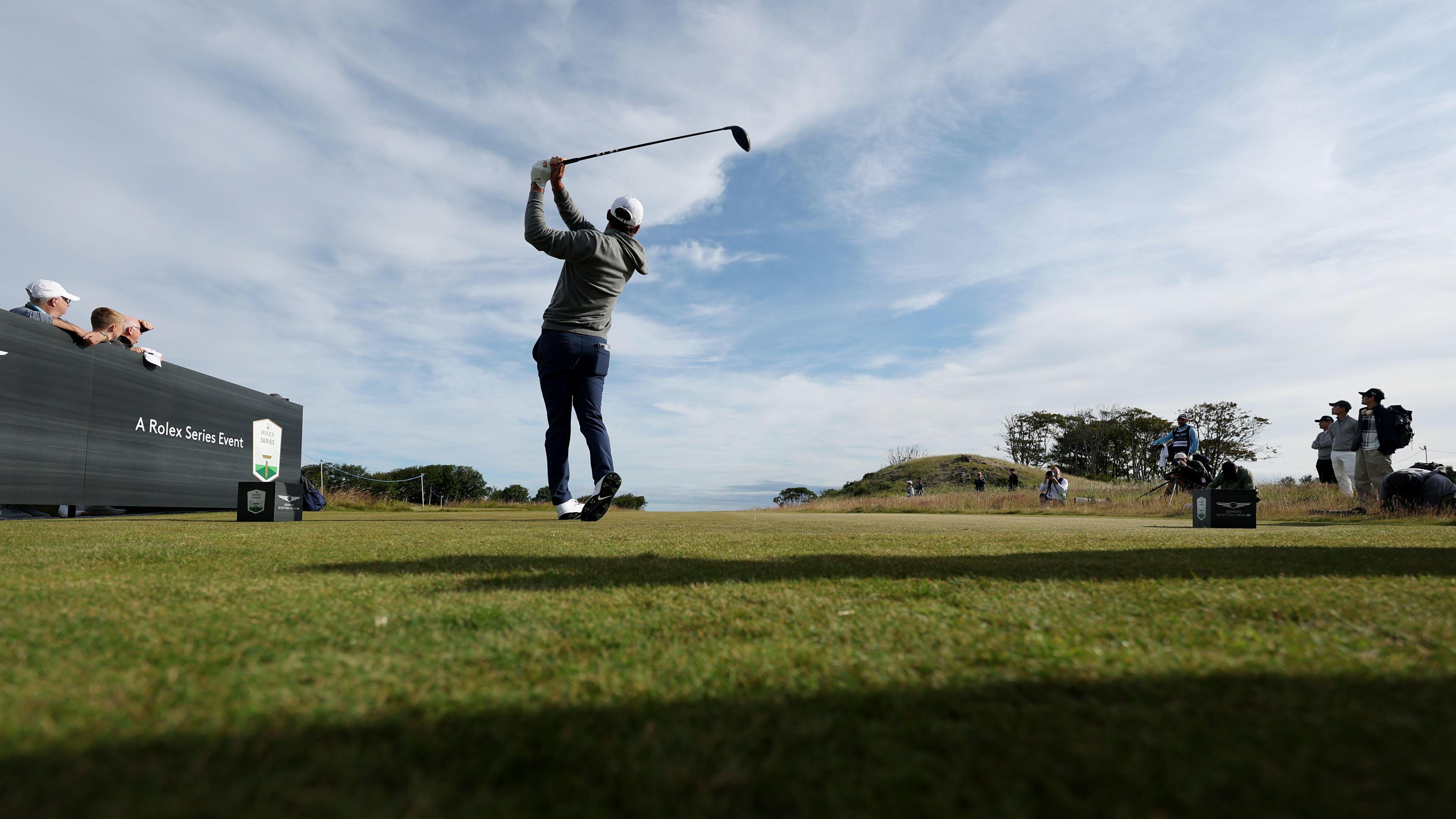 Scottie Scheffler pictured holding his driver aloft after teeing off. A small number of fans on either side follow the flight of the ball. Scheffler is wearing a grey hoodie, navy trousers, a white hat and white shoes.