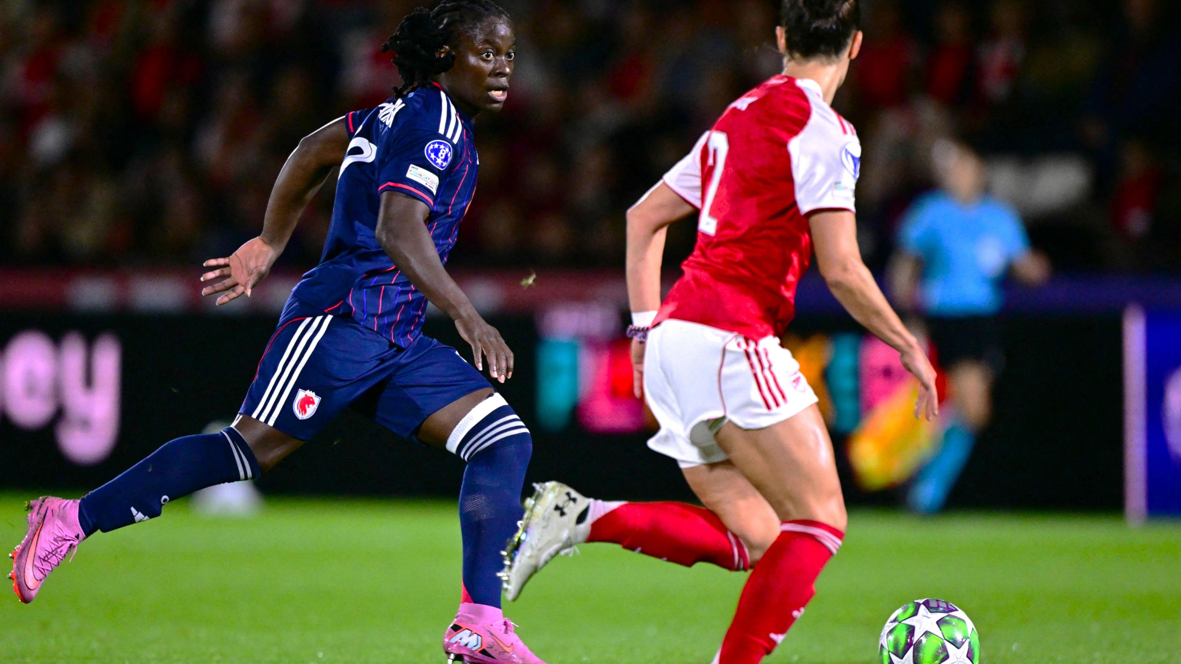 Tabitha Chawinga in all dark blue kit and pink boots is pictured mid-stride running with a football which has a star motif on it during a match between Lyon and Arsenal in the Uefa Women's Champions League. Chawinga has her head up and is looking at an opponent who is seen from behind in red and white kit coming to challenge her
