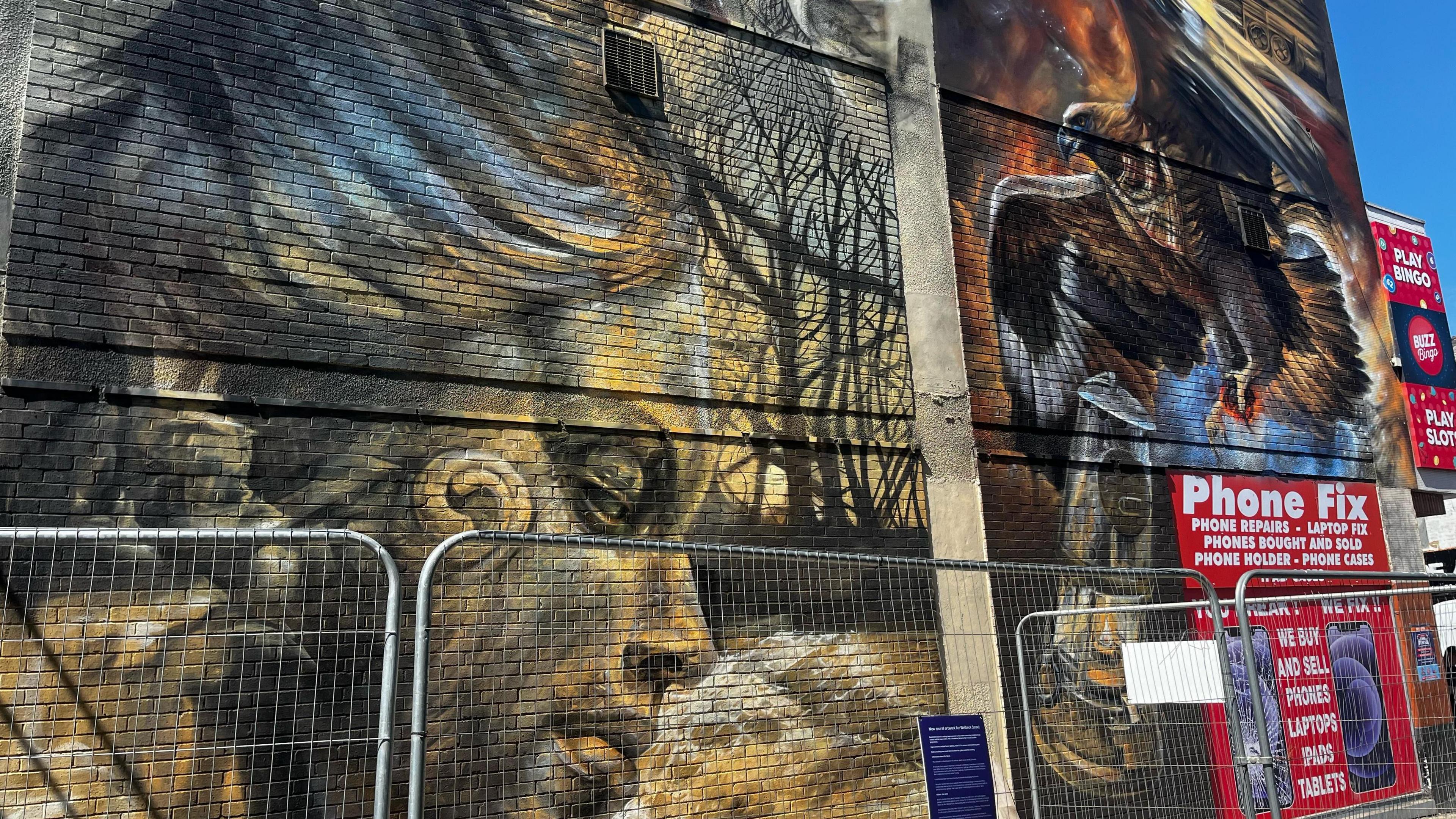 A mural on the side of a concrete wall, on the left a design of the right side of a statue's face coloured in yellow and grey, on the right an image of an eagle with wings spread holding an oil lamp. There is a phone repair advertisement in the bottom right corner and scaffolding in front of the mural.